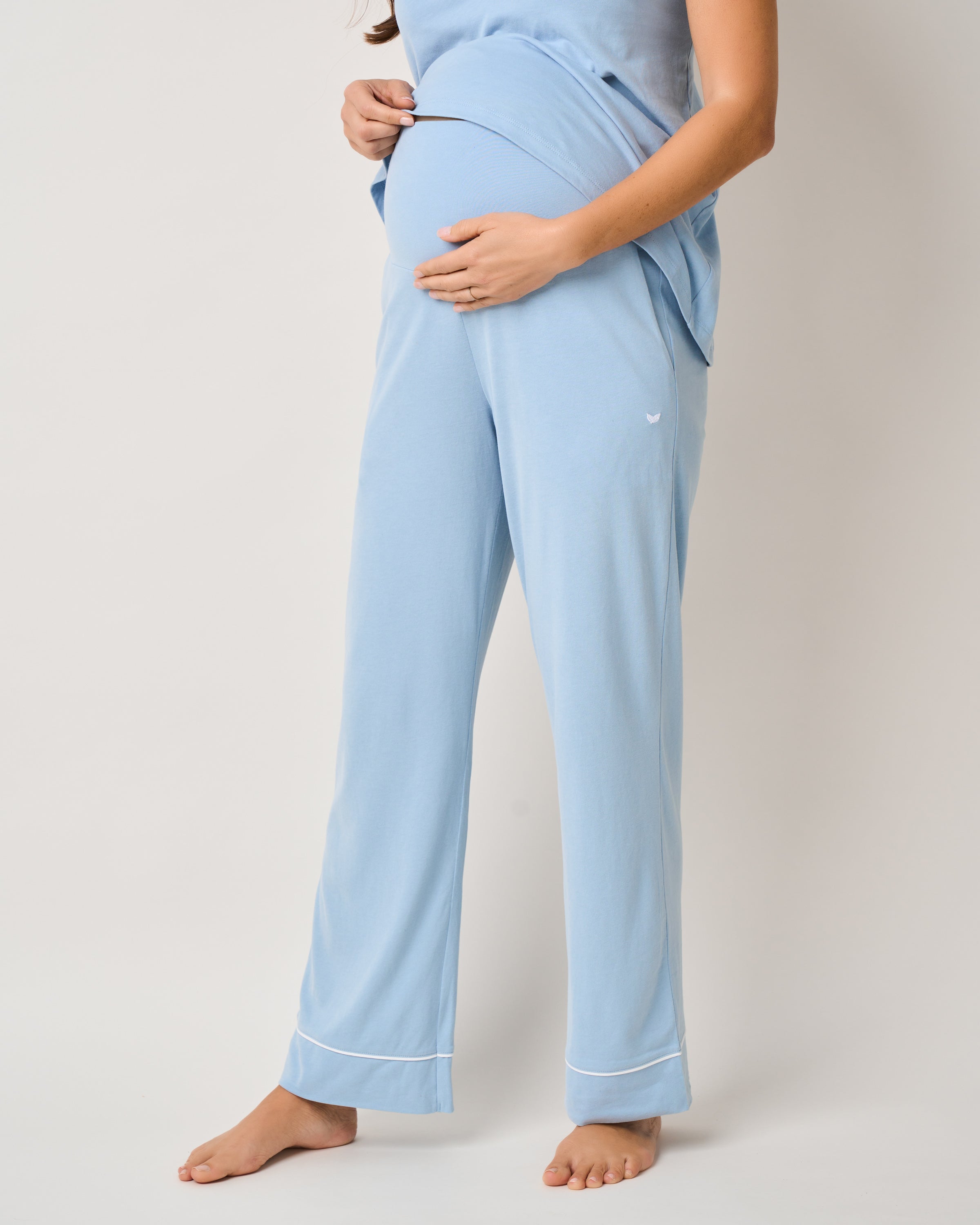 A pregnant woman in Petite Plumes Womens Pima Maternity Pants in Periwinkle stands barefoot, gently cradling her belly with an over-the-belly support band, set against a plain white background.