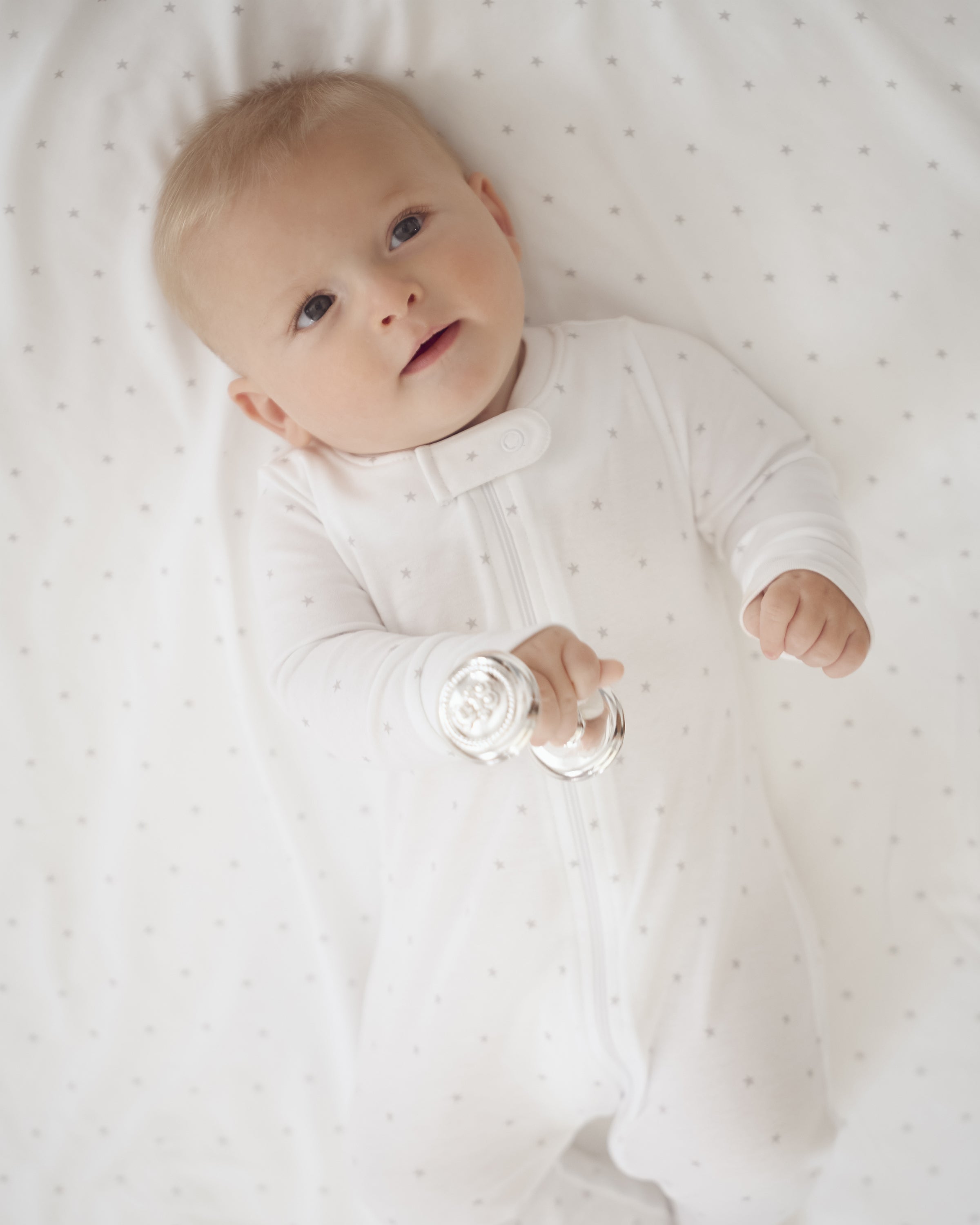 A baby wearing a soft white onesie lies on a Petite Plume Pima Baby Blanket in Grey Stars, holding a silver rattle, with Peruvian Pima Cotton ensuring comfort as the baby looks upward with a gentle expression.