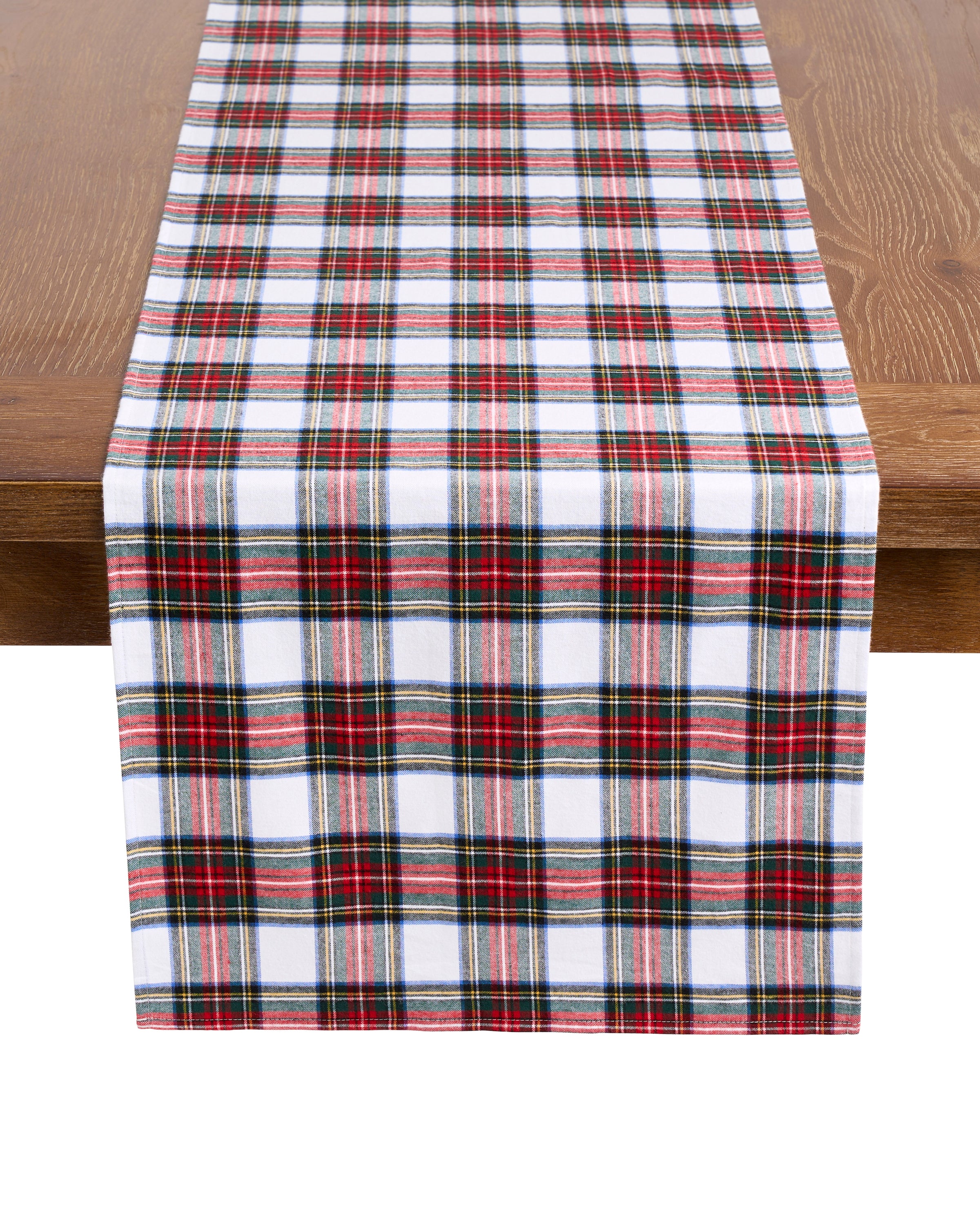 A wooden table features the Petite Plume Signature Twill Table Runner in Balmoral Tartan, with red, green, white, and blue plaid, effortlessly enhancing a festive tablescape.