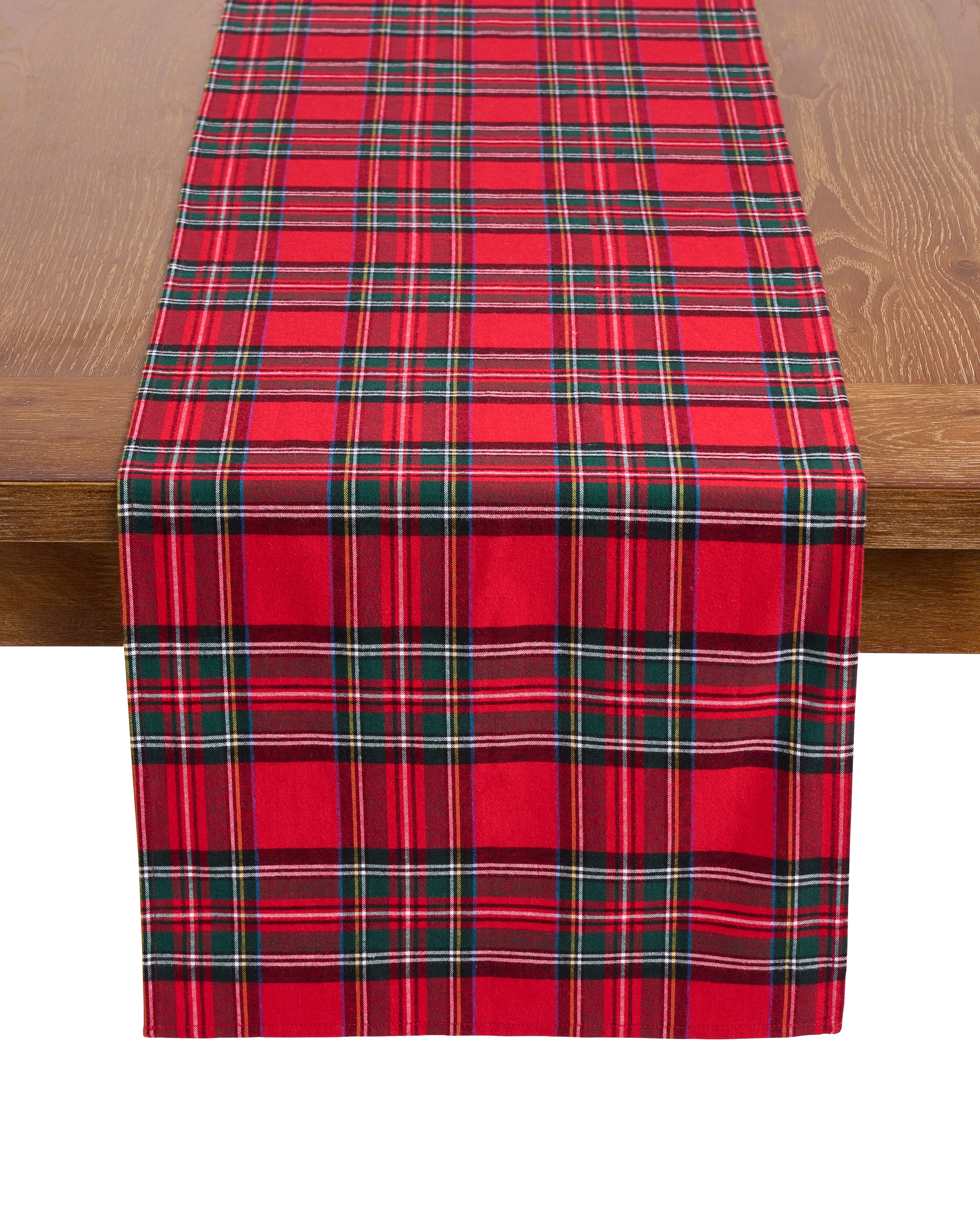 Petite Plumes Signature Twill Table Runner in Imperial Tartan, with its red, green, and white plaid design, graces a wooden table. This festive piece enhances any occasion and beautifully complements other linens.