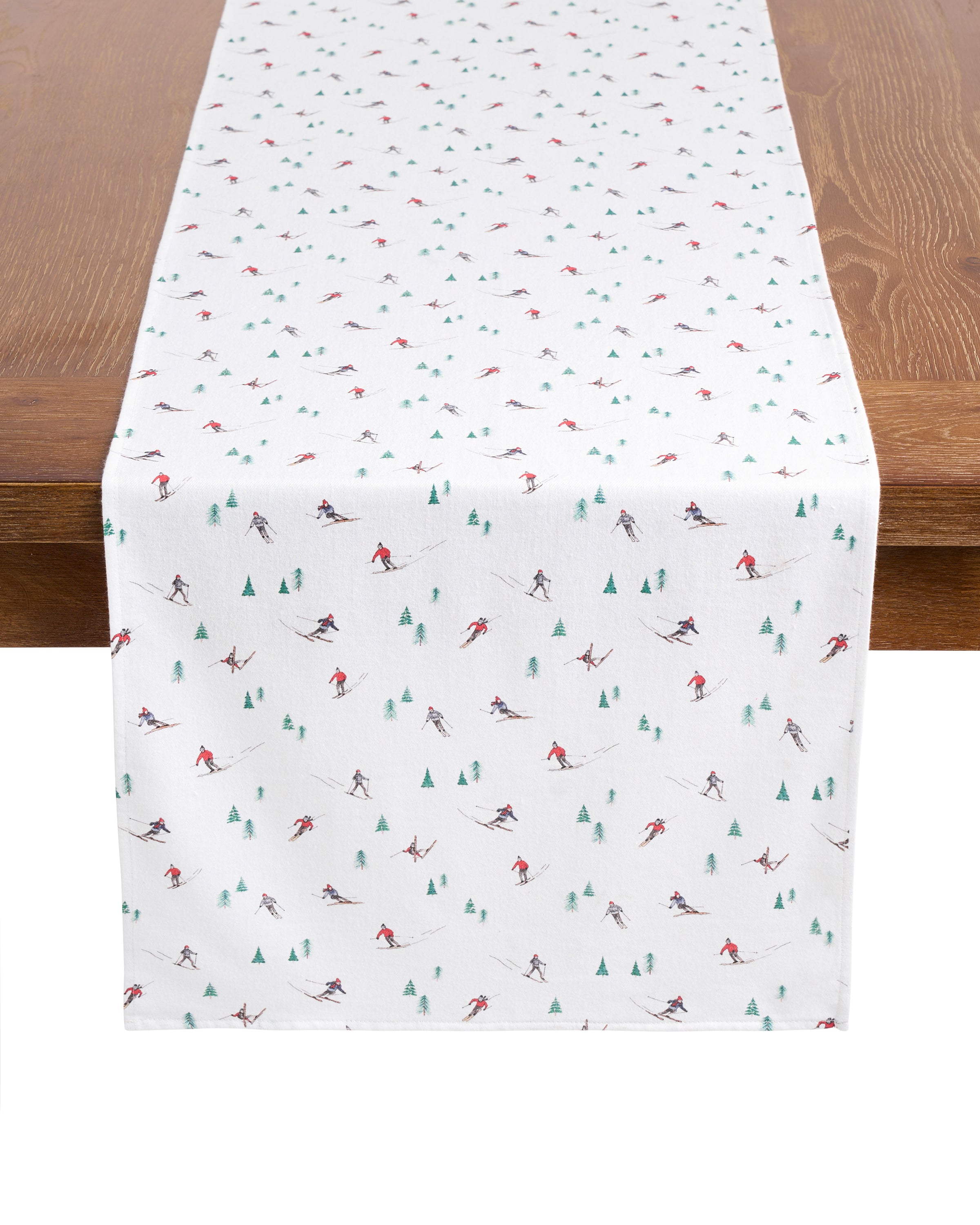 A wooden table features Petite Plumes Signature Twill Table Runner in Après Ski, displaying a festive design with small skiers and trees in cheerful red, green, and gray.