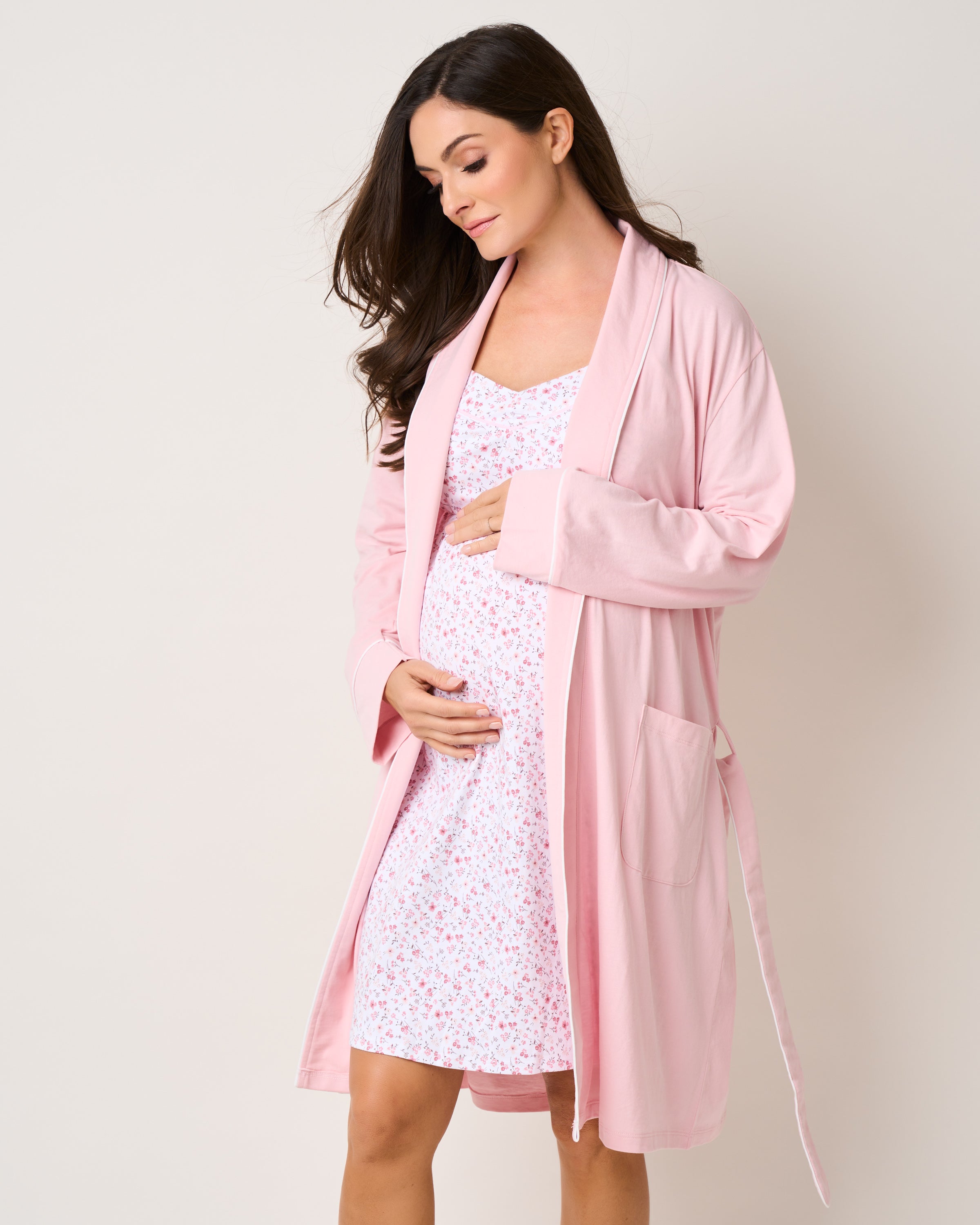 A pregnant woman gently cradles her belly, wrapped in a pink robe over Petite Plumes Womens Pima Maternity Nightgown in Dorset Floral. Her long dark hair cascades over her shoulders as she looks downward serenely against a plain, light-colored background.