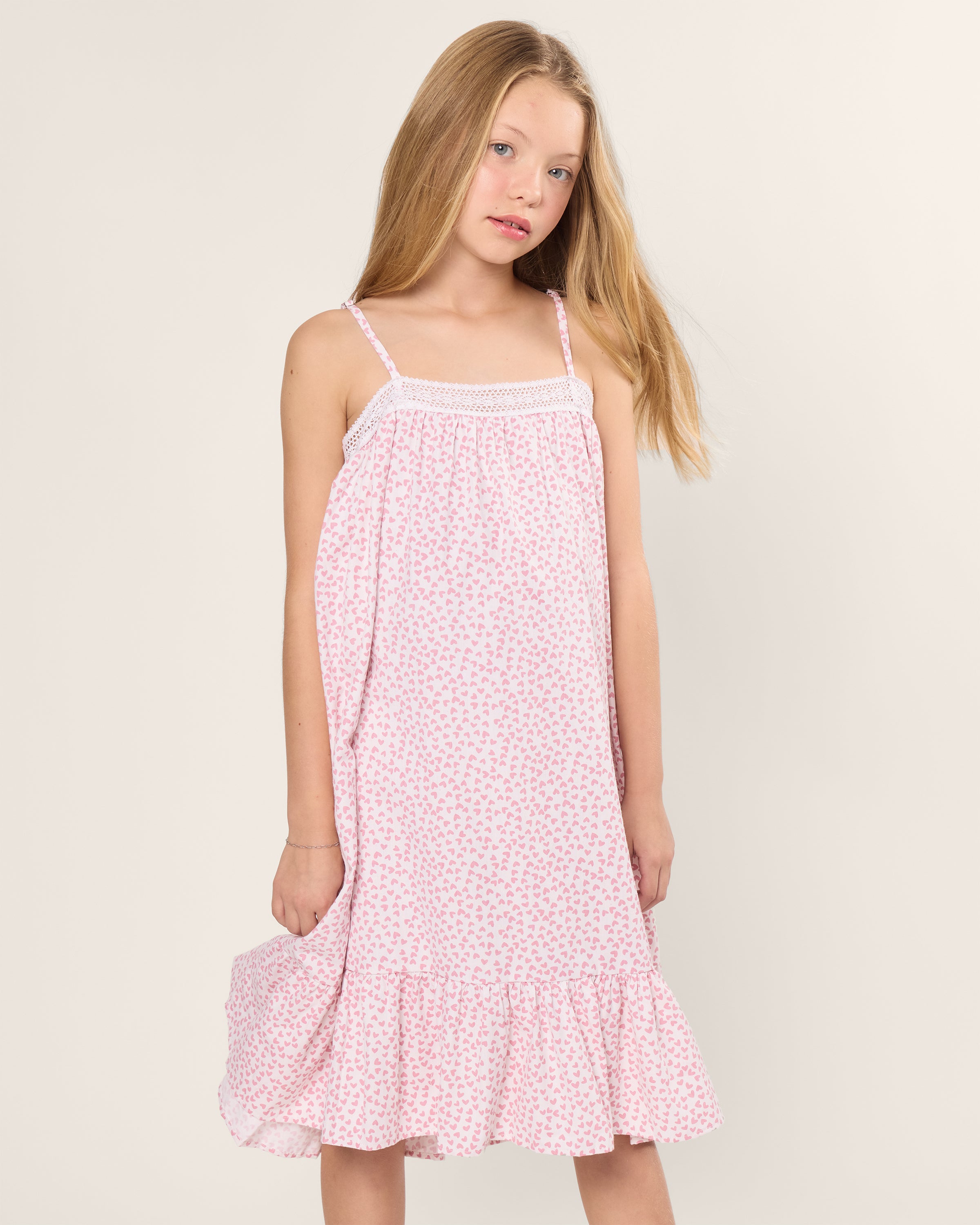 A young girl in a Girls Twill Lily Nightgown in Sweethearts by Petite Plume stands against a plain background. Her long blonde hair cascades freely, exuding relaxed comfort reminiscent of luxurious yarn-dyed cotton sleepwear.
