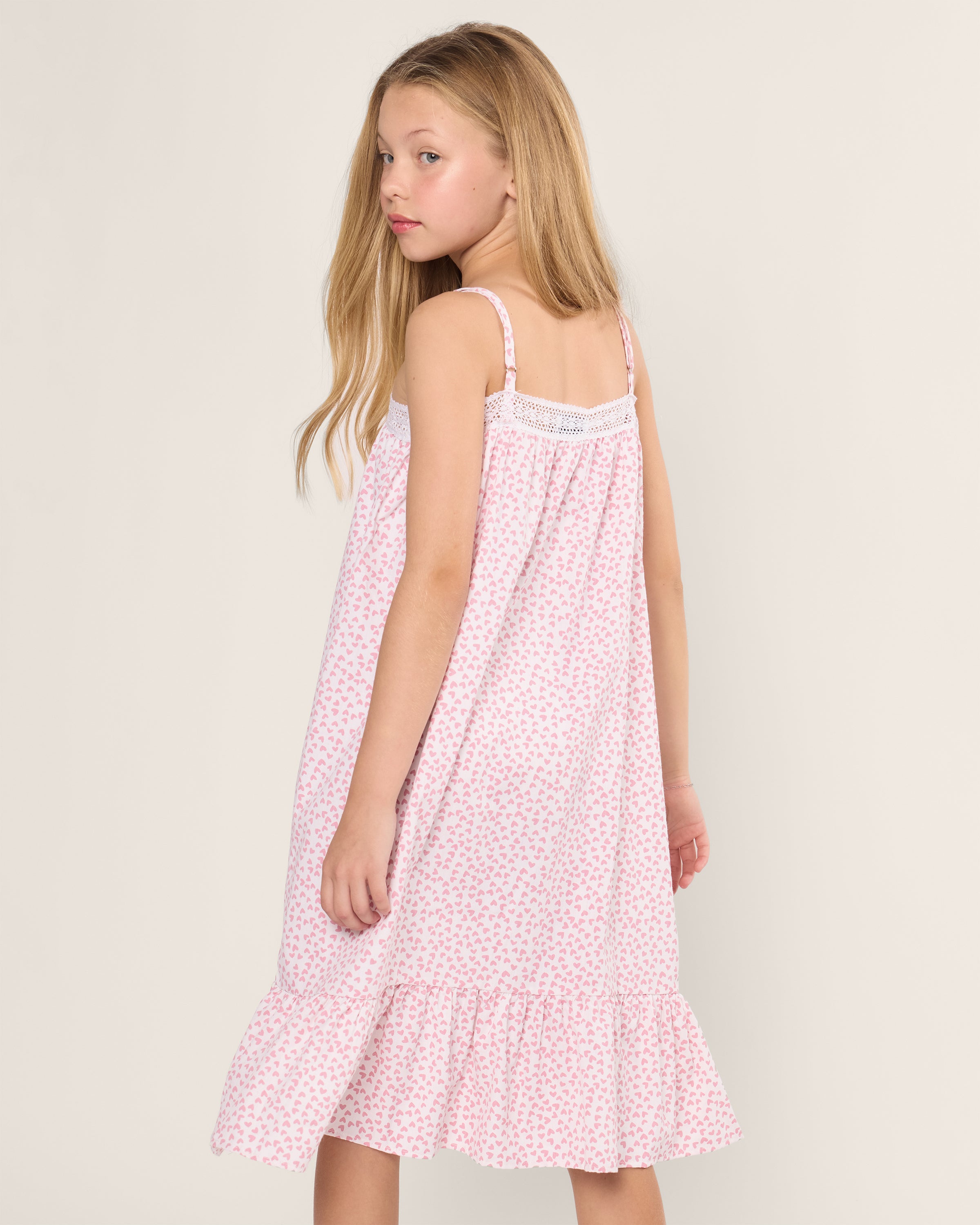 A young girl with long blonde hair wears a pink and white floral dress made from yarn-dyed cotton, with delicate lace details. She stands turned, looking over her shoulder against a plain light background, echoing the softness of Petite Plumes Twill Lily Nightgown in Sweethearts.