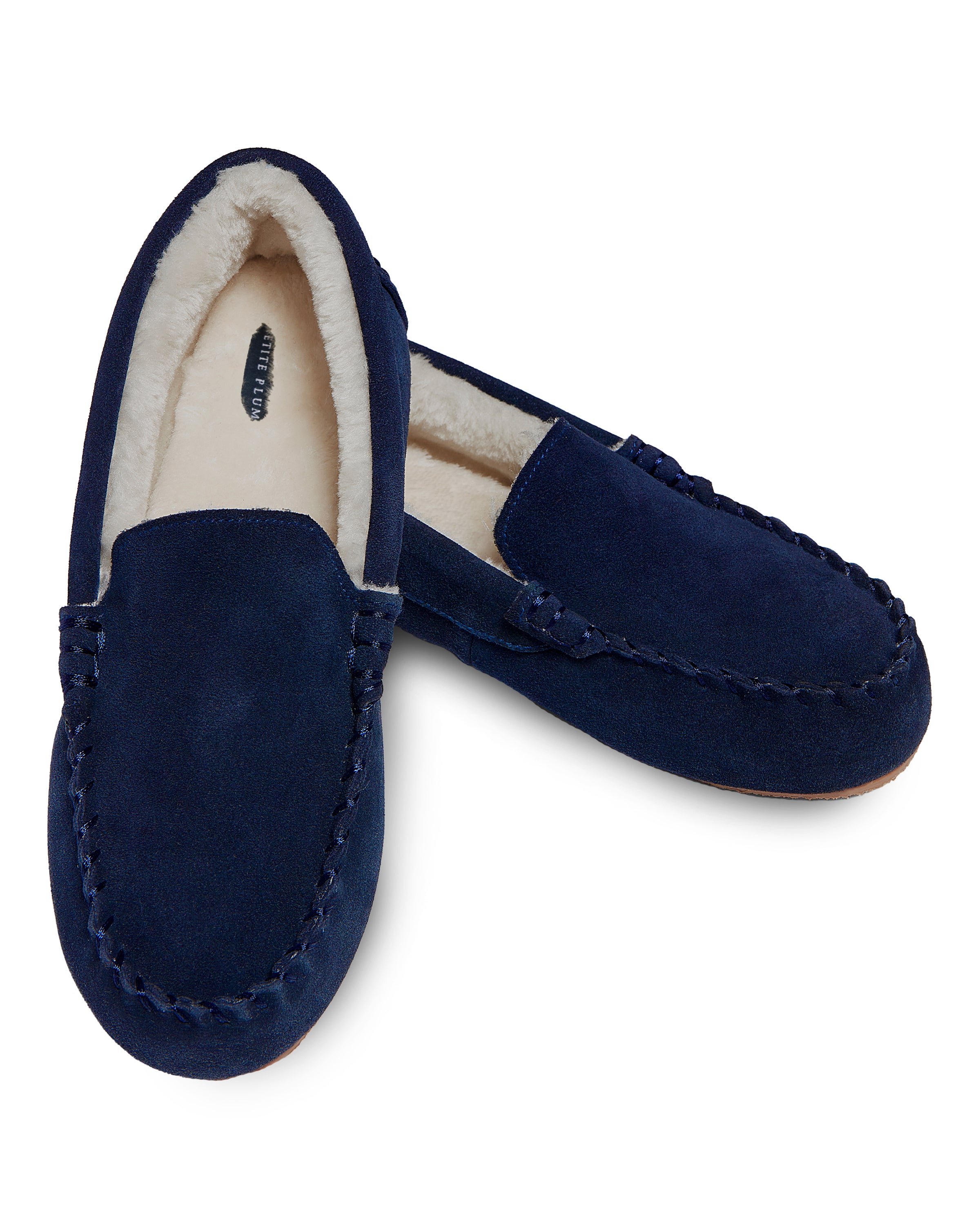 Mens suede slippers in navy by Petite Plume feature a plush, ultra-soft faux fur lining. One slipper is slightly turned outward, showing intricate stitching and a cozy interior. The light brown outsole completes this comfortable design.
