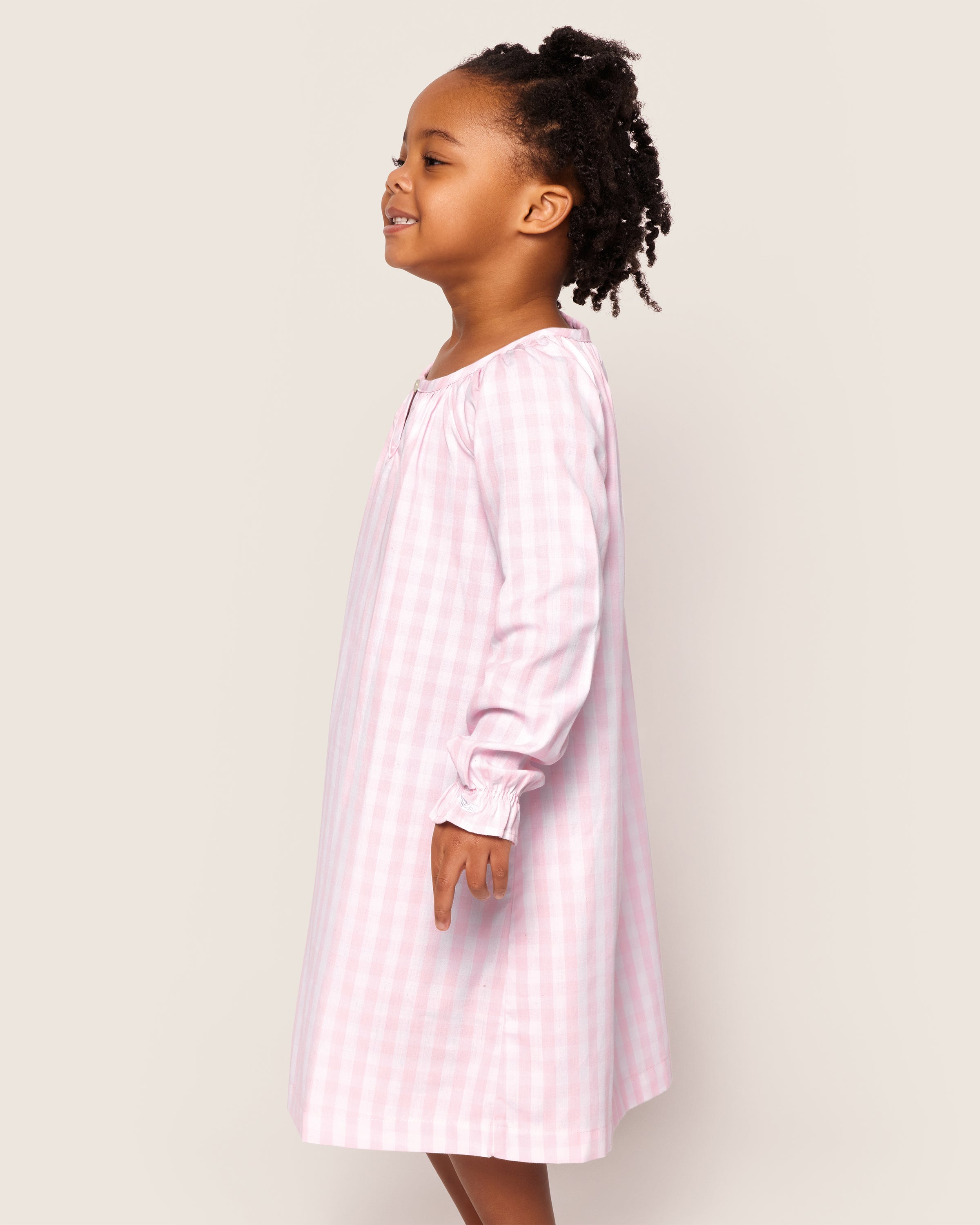 A young girl smiles in profile, wearing Petite Plumes Girls Twill Delphine Nightgown in pink gingham. Her hair is styled in multiple small braids against a plain, light-colored background.
