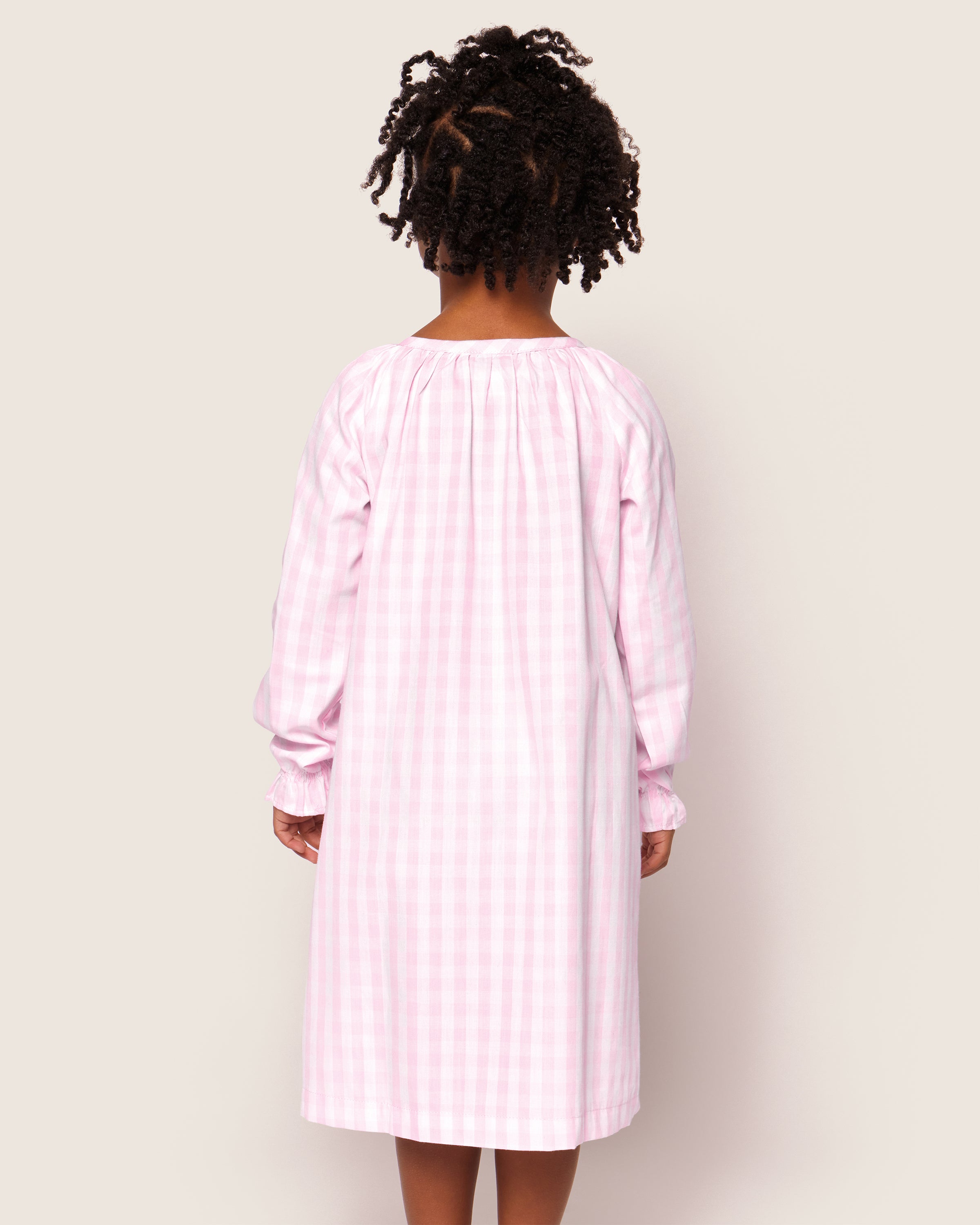A curly-haired child is seen from behind against a plain background, wearing Petite Plumes Girls Twill Delphine Nightgown in Pink Gingham.
