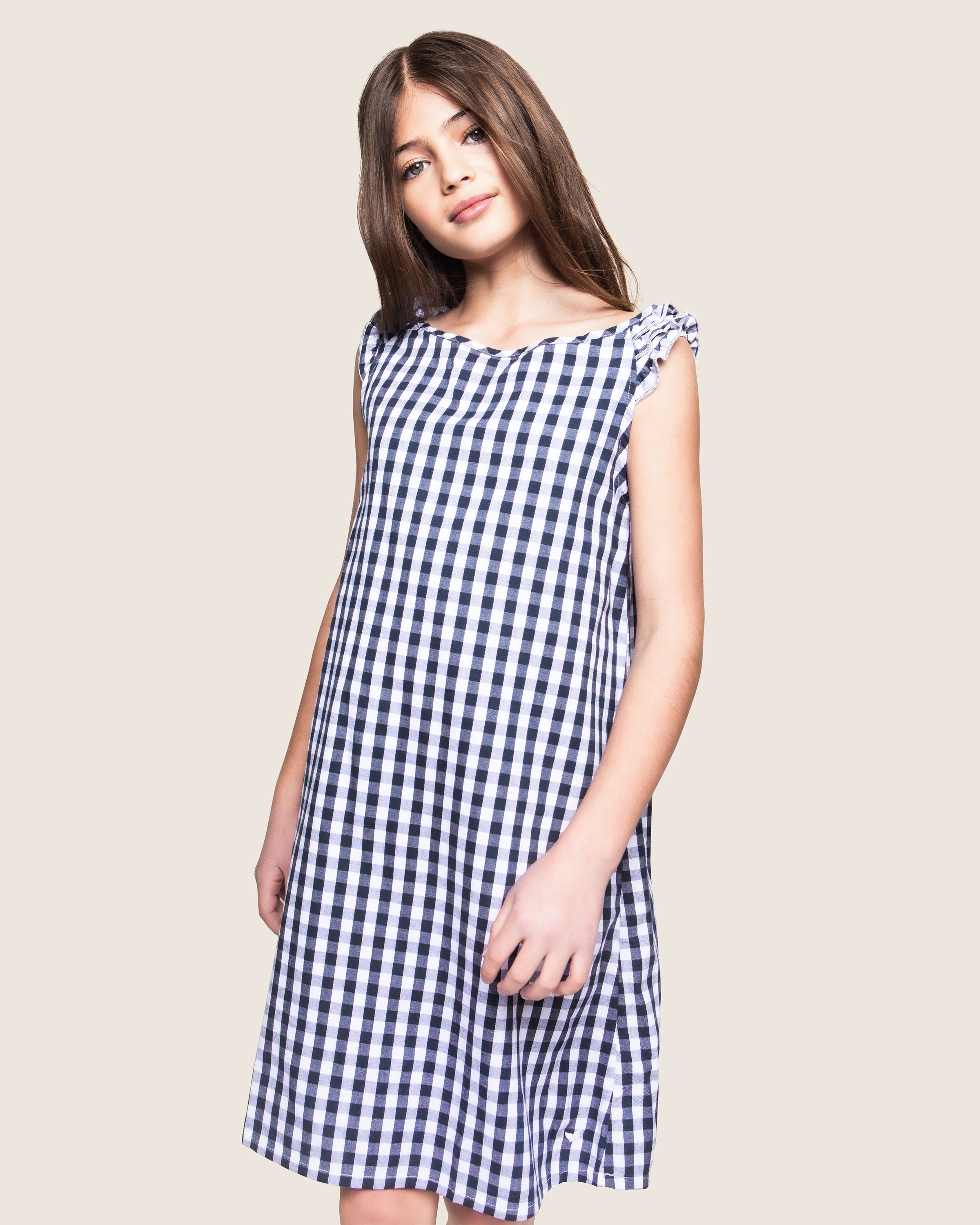 A young girl with long brown hair wears Petite Plumes Girls Twill Amelie Nightgown in navy gingham. She stands with a slight lean, looking directly at the camera with a neutral expression. The background is plain off-white.