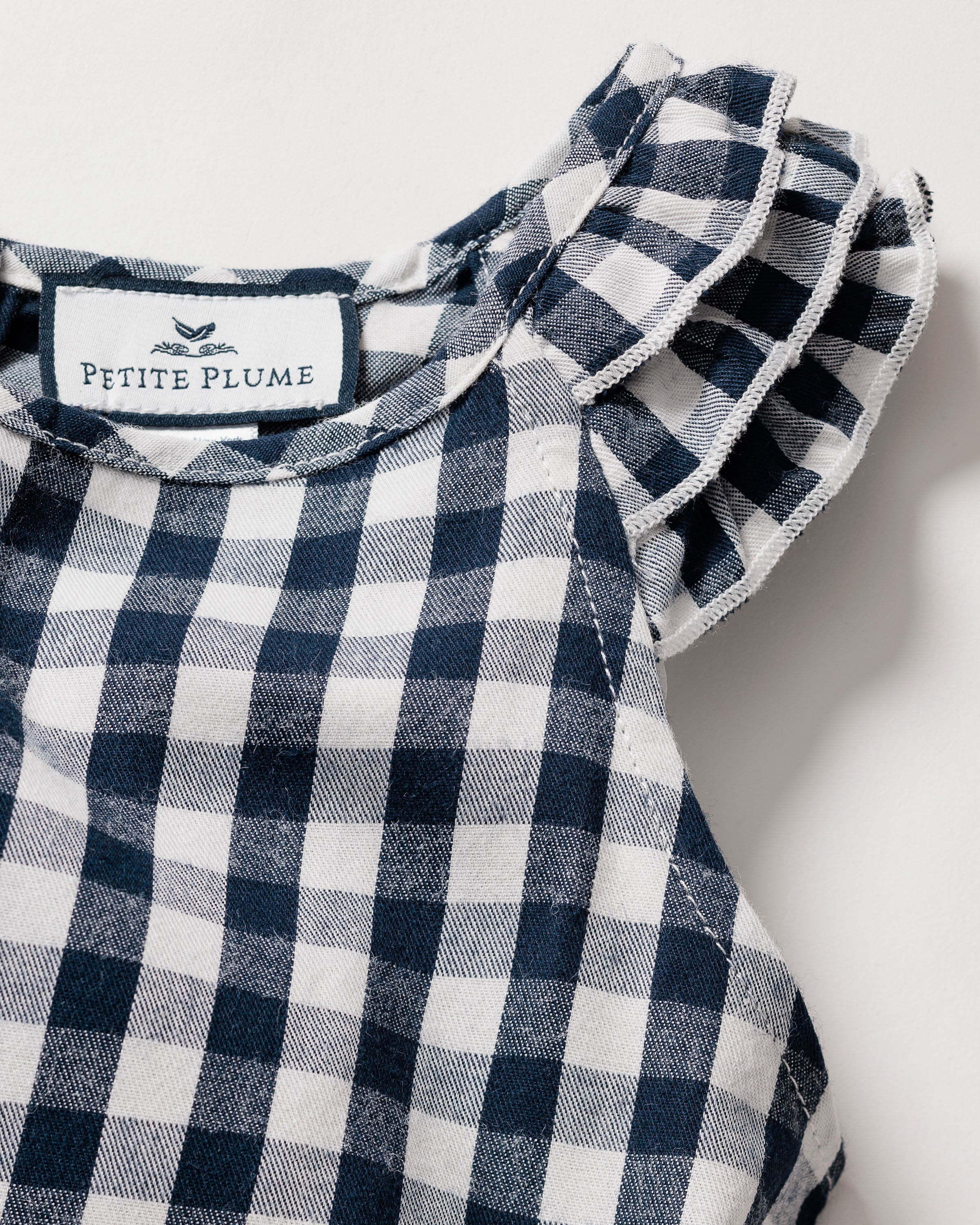 Close-up of a navy gingham print dress with ruffled sleeves. This lightweight twill nightgown has a fabric label at the neckline with Petite Plume and a small bird illustration, featuring a soft, textured appearance.