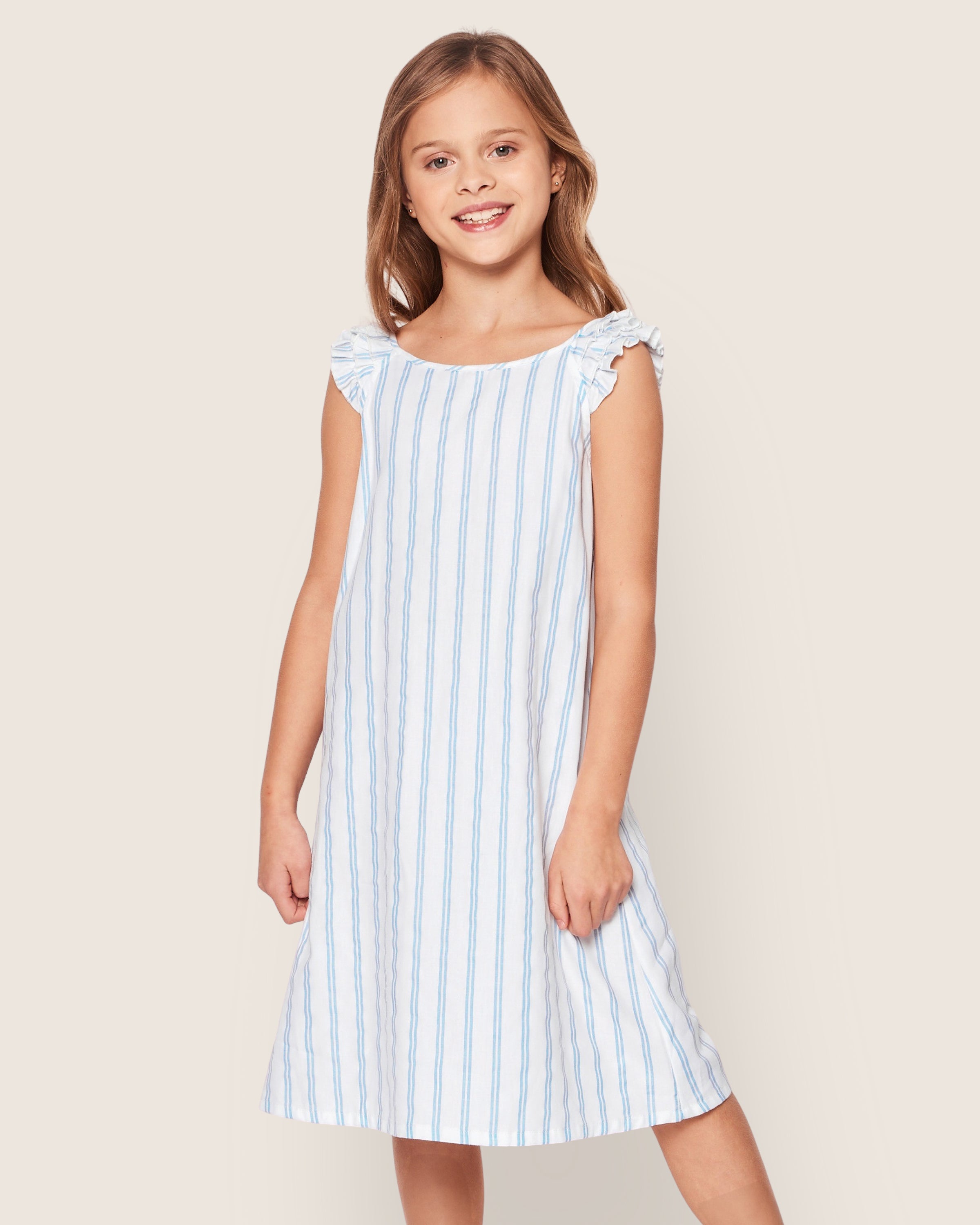 A young girl smiles in Petite Plumes Girls Twill Amelie Nightgown in Periwinkle Stripe, featuring a light blue and white design with ruffled shoulders. She stands against a plain, light background with her hair down and posture relaxed.