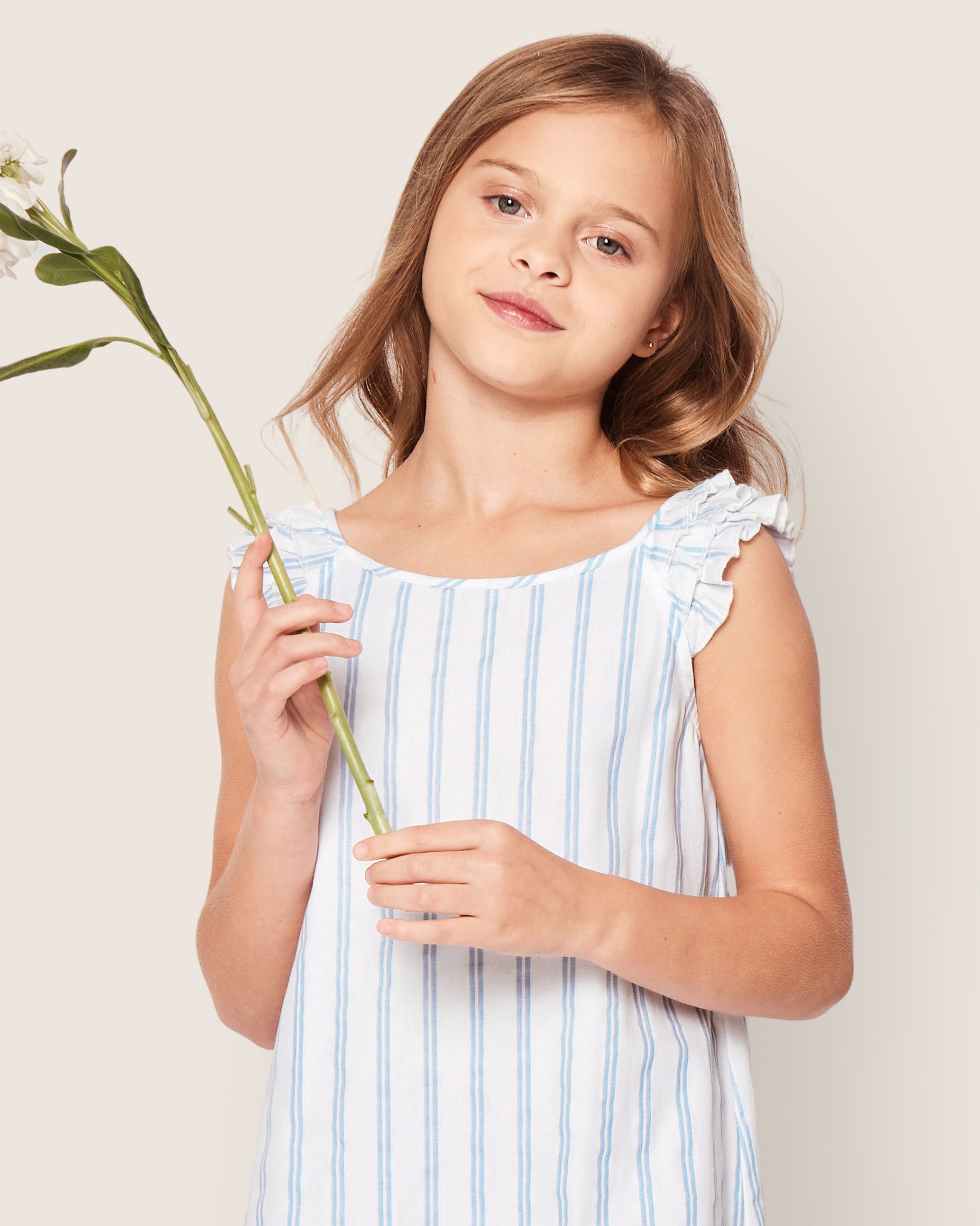 A young girl in a Petite Plume Girls Twill Amelie Nightgown in Periwinkle Stripe holds a white flower. Her long, light brown hair frames her relaxed expression as she stands against a simple, light background.