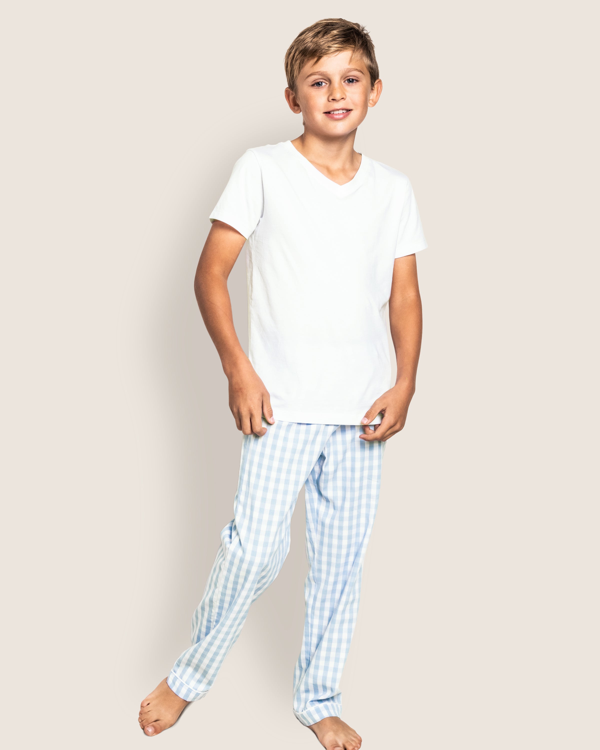 A child stands against a plain background, smiling in their luxurious sleepwear from Petite Plume. They wear a white T-shirt and Kids Twill Pajama Pants in Light Blue Gingham made from yarn-dyed cotton, with bare feet.