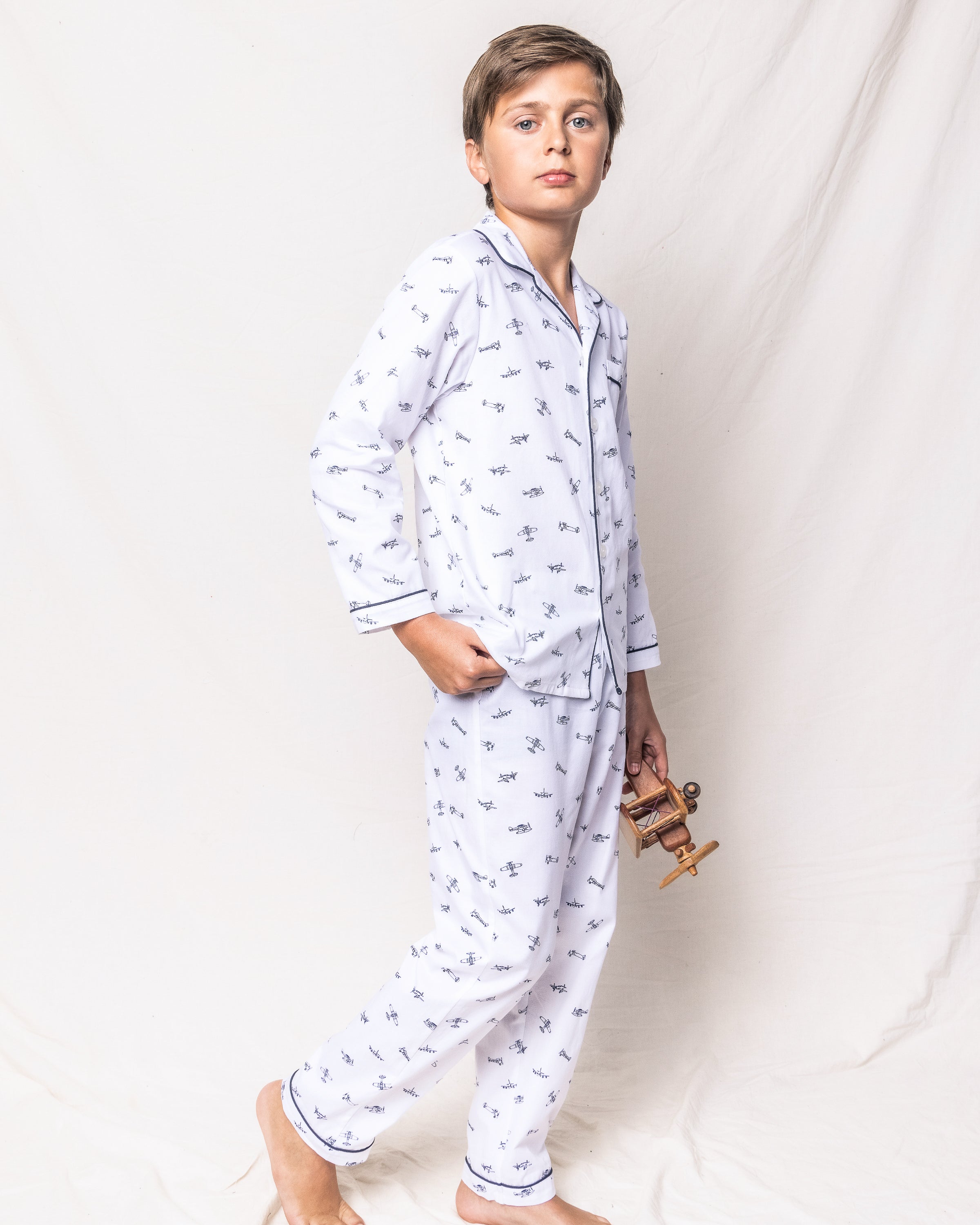 A boy in Petite Plumes Kids Twill Pajama Set with Par Avion print stands against a plain backdrop, holding a small wooden airplane.