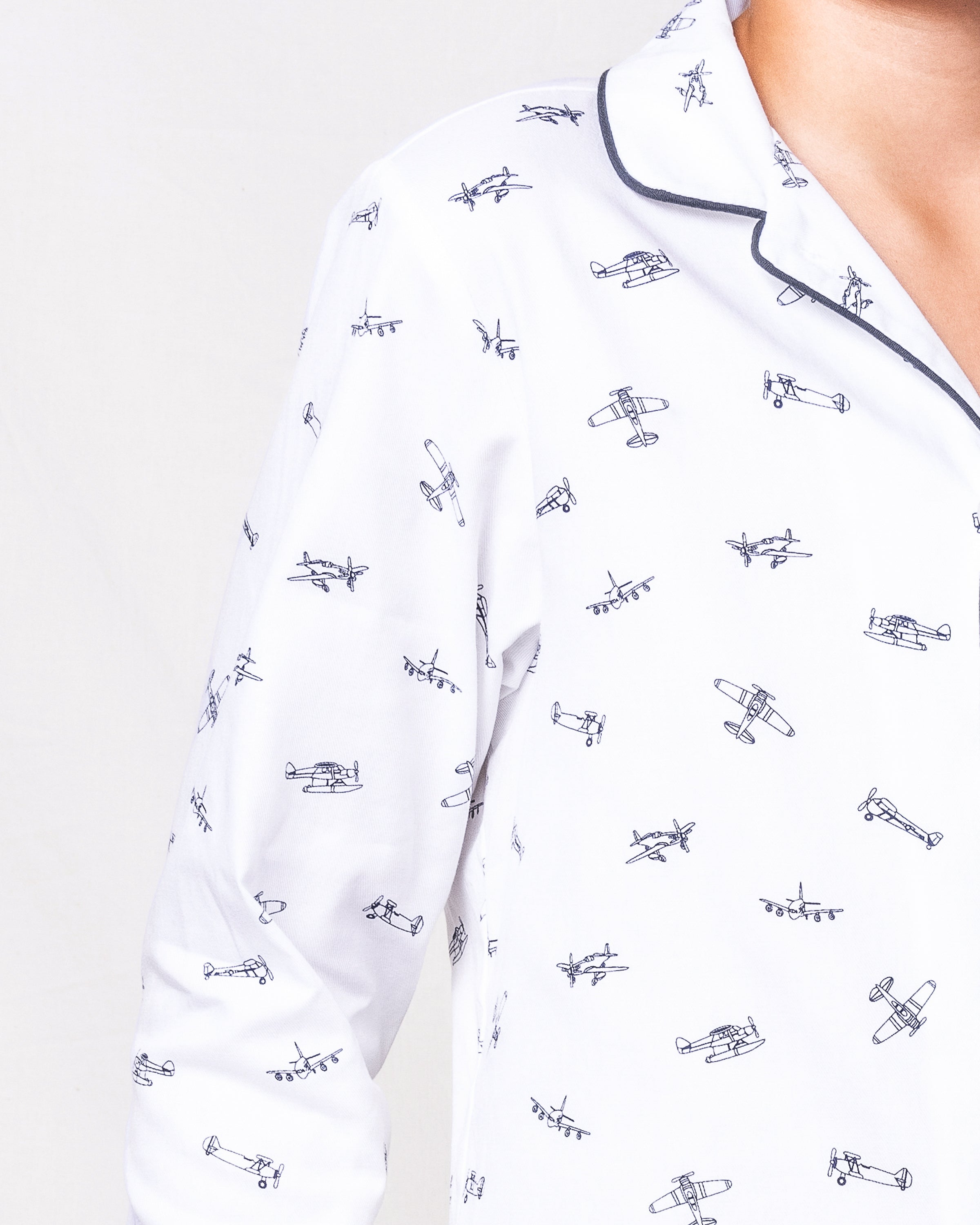 Close-up of a person wearing Petite Plumes Kids Twill Pajama Set in Par Avion, showing vintage airplane prints. The white top features a collar with dark piping, highlighting luxurious sleepwear against a plain background with only the right side and one shoulder visible.