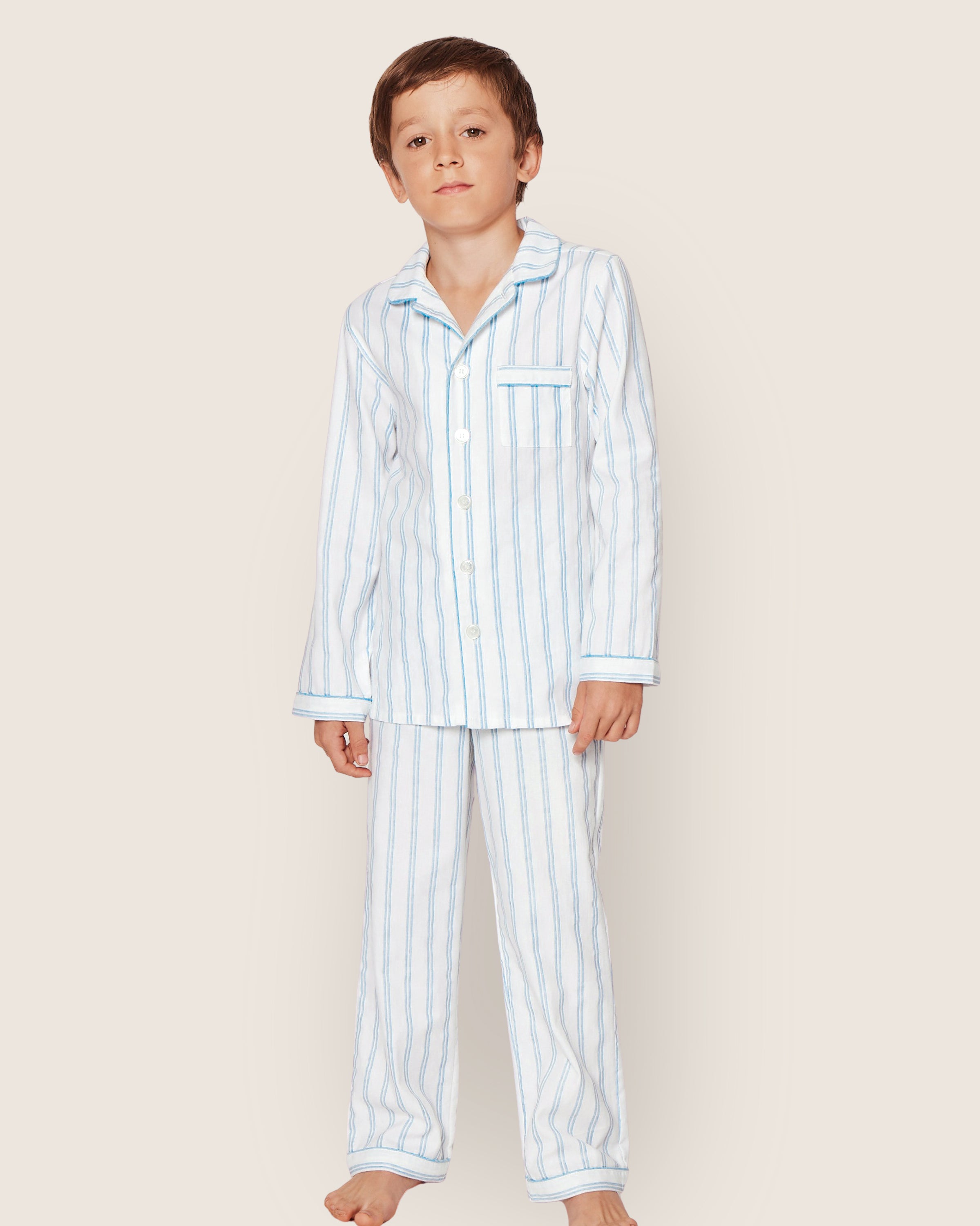 A young boy stands barefoot in the Petite Plume Kids Twill Pajama Set—periwinkle and white stripes, featuring a button-up shirt and matching pants—contrasting against a light plain background.