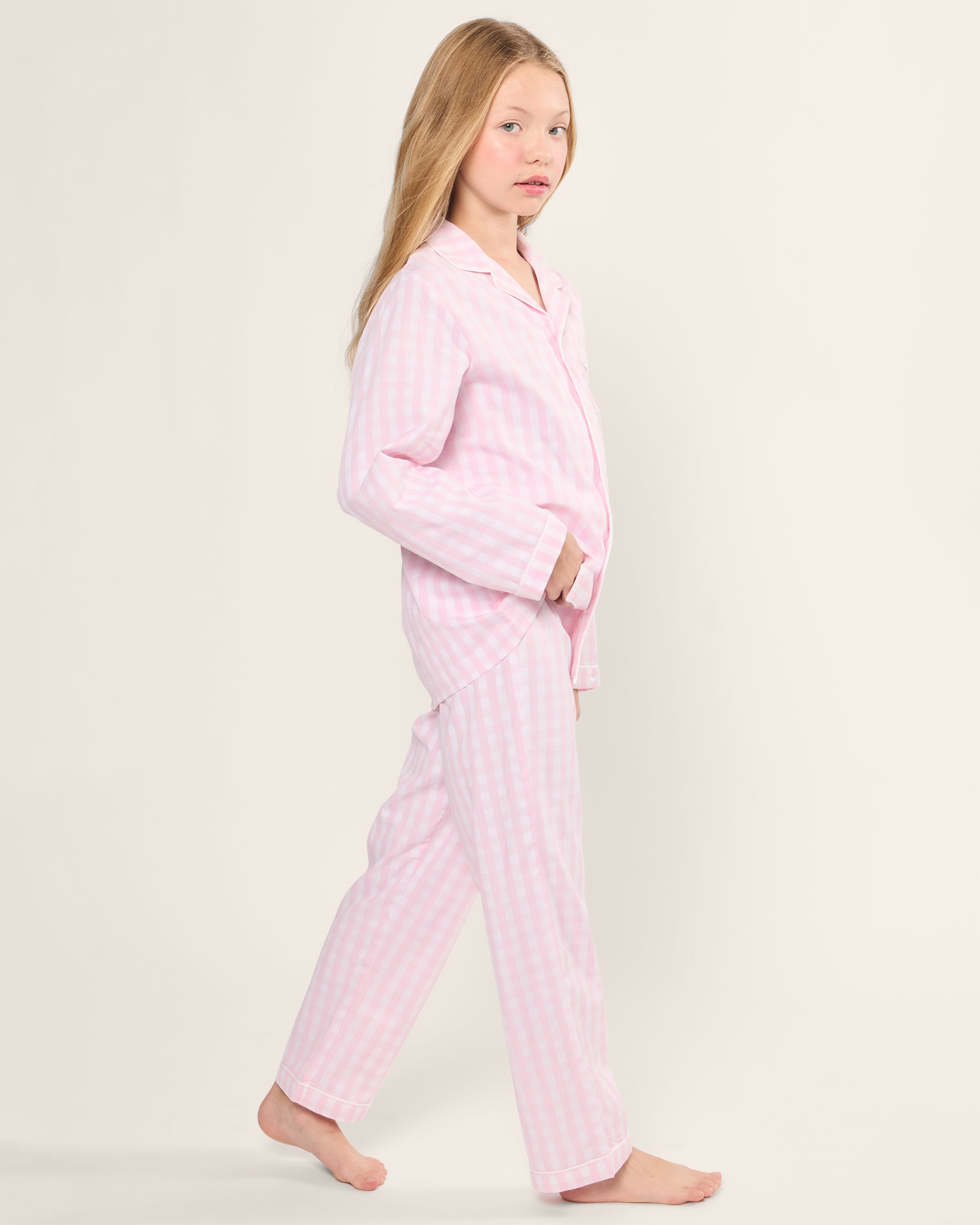 A young girl with long blonde hair walks barefoot across a plain background, dressed in a flame retardant Petite Plume Kids Twill Pajama Set in Pink Gingham made from yarn-dyed cotton.