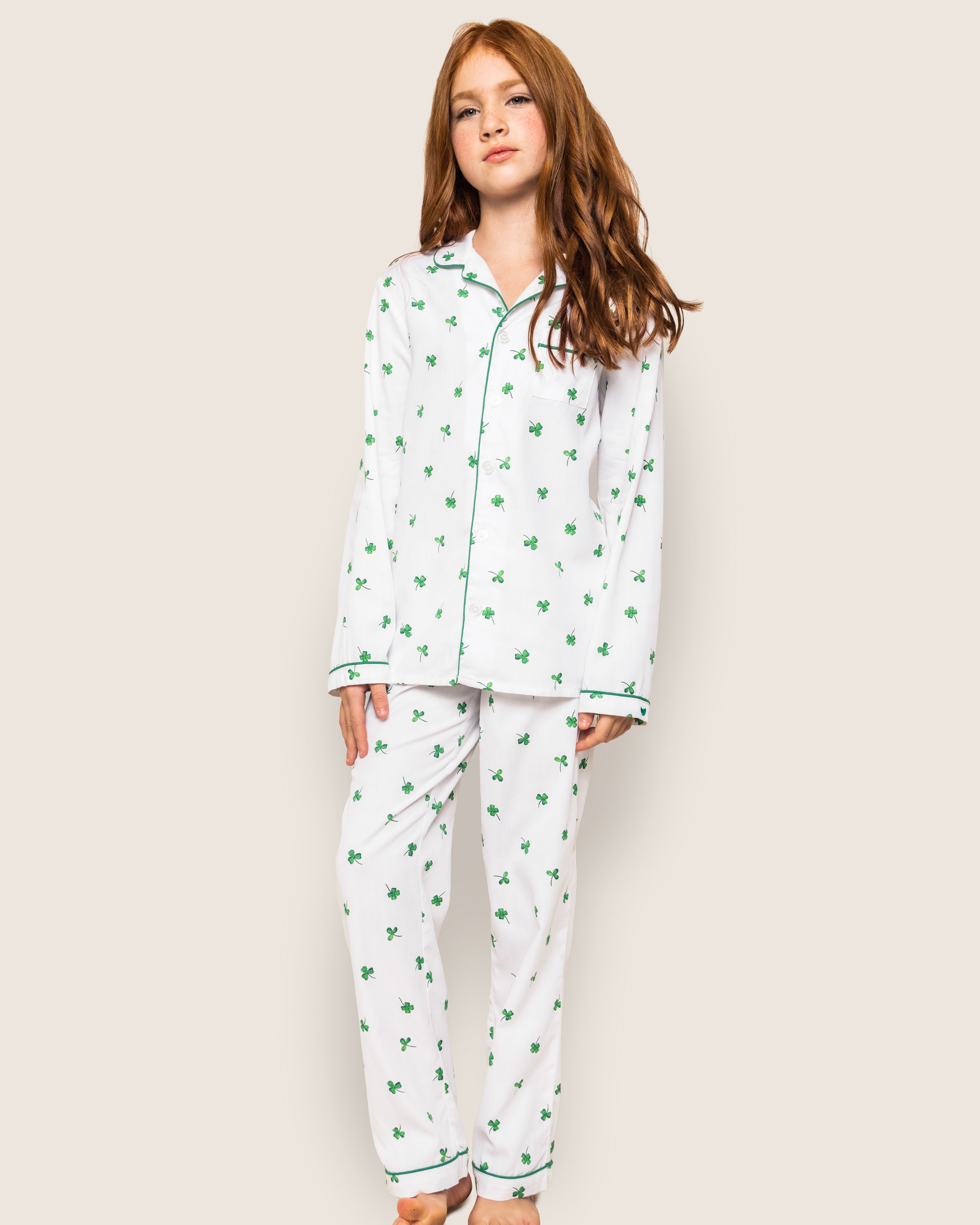 A young girl with long red hair stands against a plain background in the Kids Twill Pajama Set in Shamrocks from Petite Plume. She looks ahead neutrally, her hands at her sides.
