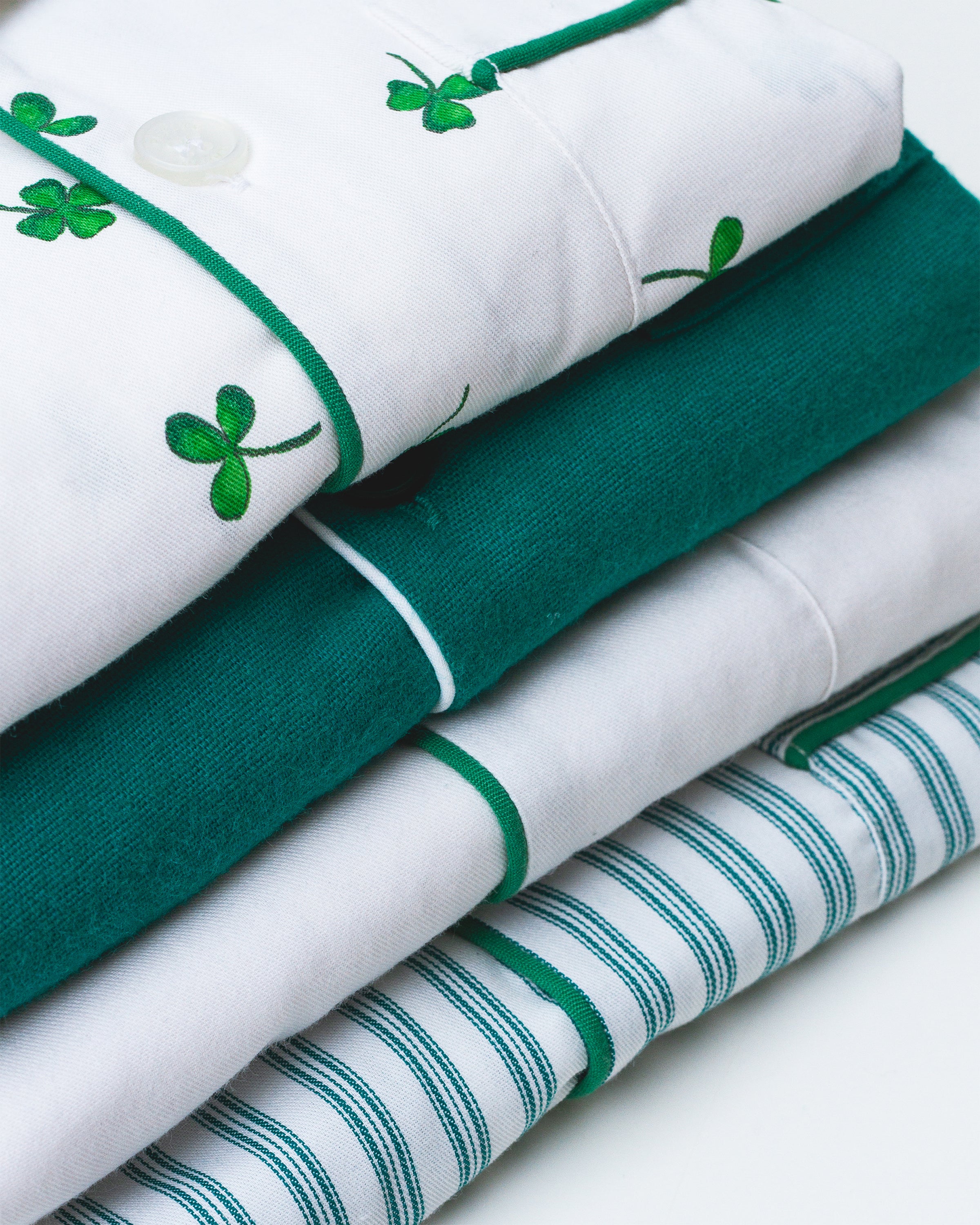 A stack of folded Kids Twill Pajama Sets in Shamrocks from Petite Plume, showcasing green clover, solid green, and green-striped designs, is neatly layered against a pristine white background.