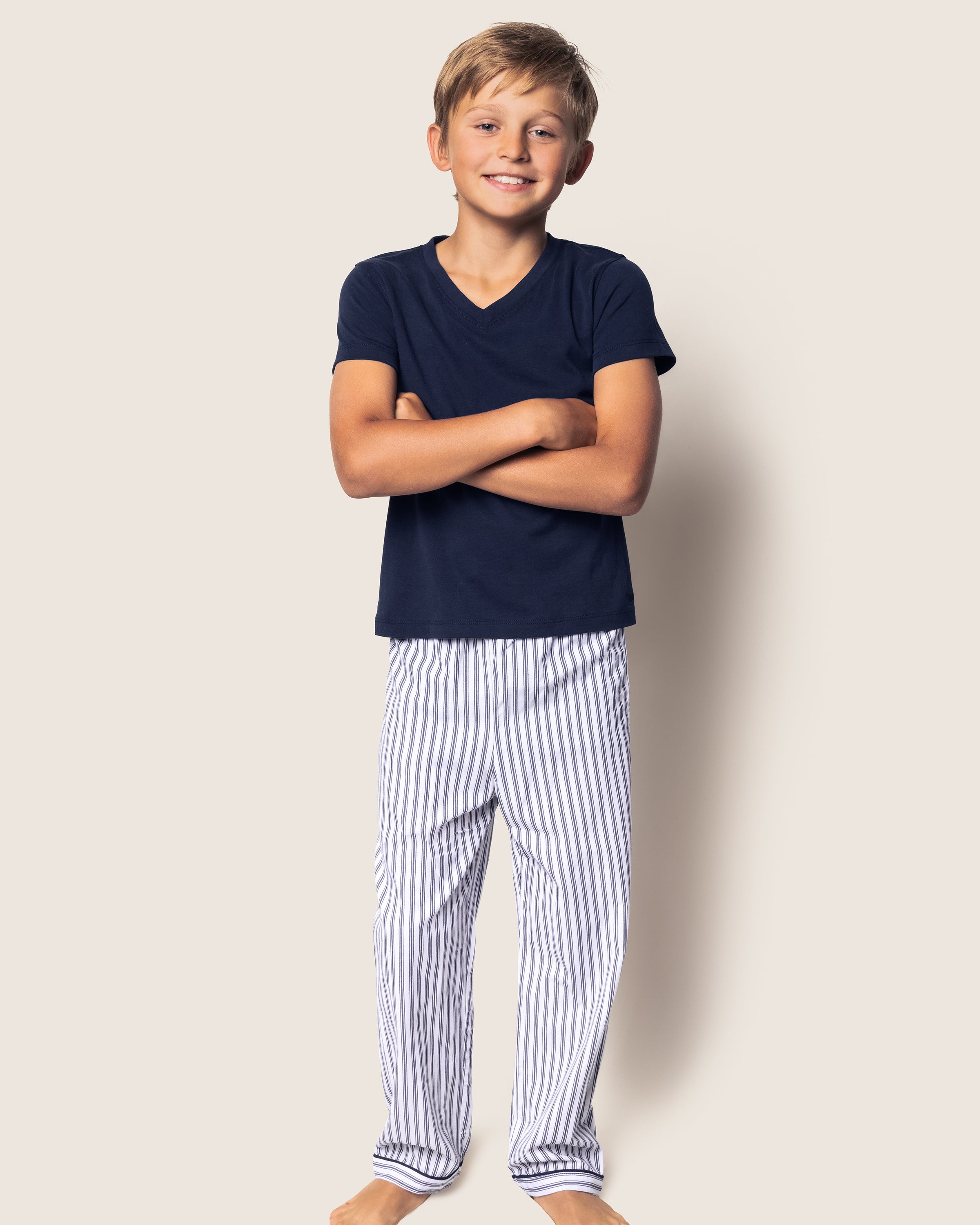 A smiling boy stands with arms crossed, proudly wearing a navy T-shirt and Petite Plumes navy twill pajama pants in French ticking. The plain off-white background highlights his charming outfit.
