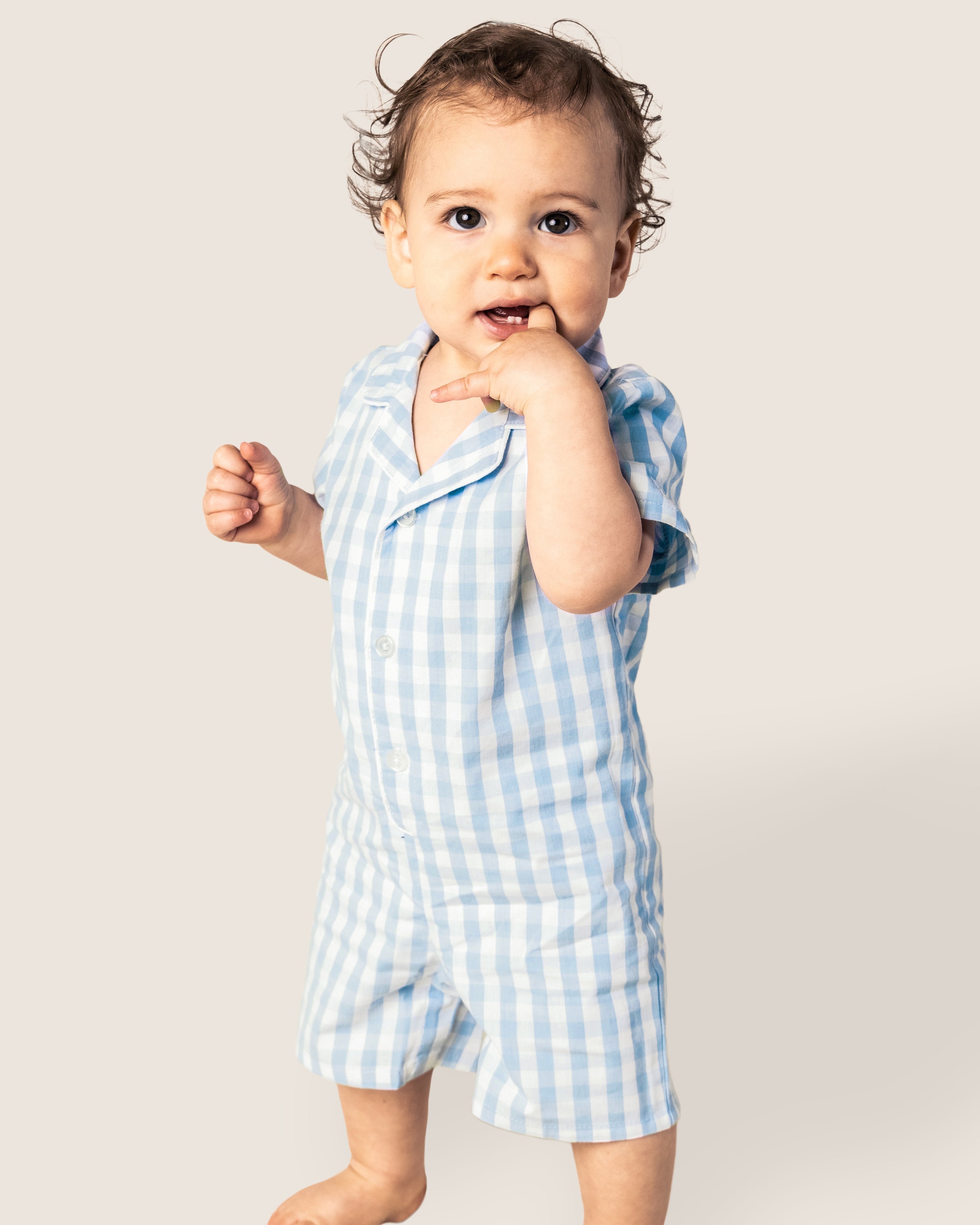 A toddler stands barefoot against a plain background, wearing Petite Plumes Babys Twill Summer Romper in light blue gingham. Made from yarn-dyed cotton, the child looks forward with one hand partially in their mouth and the other relaxed by their side.