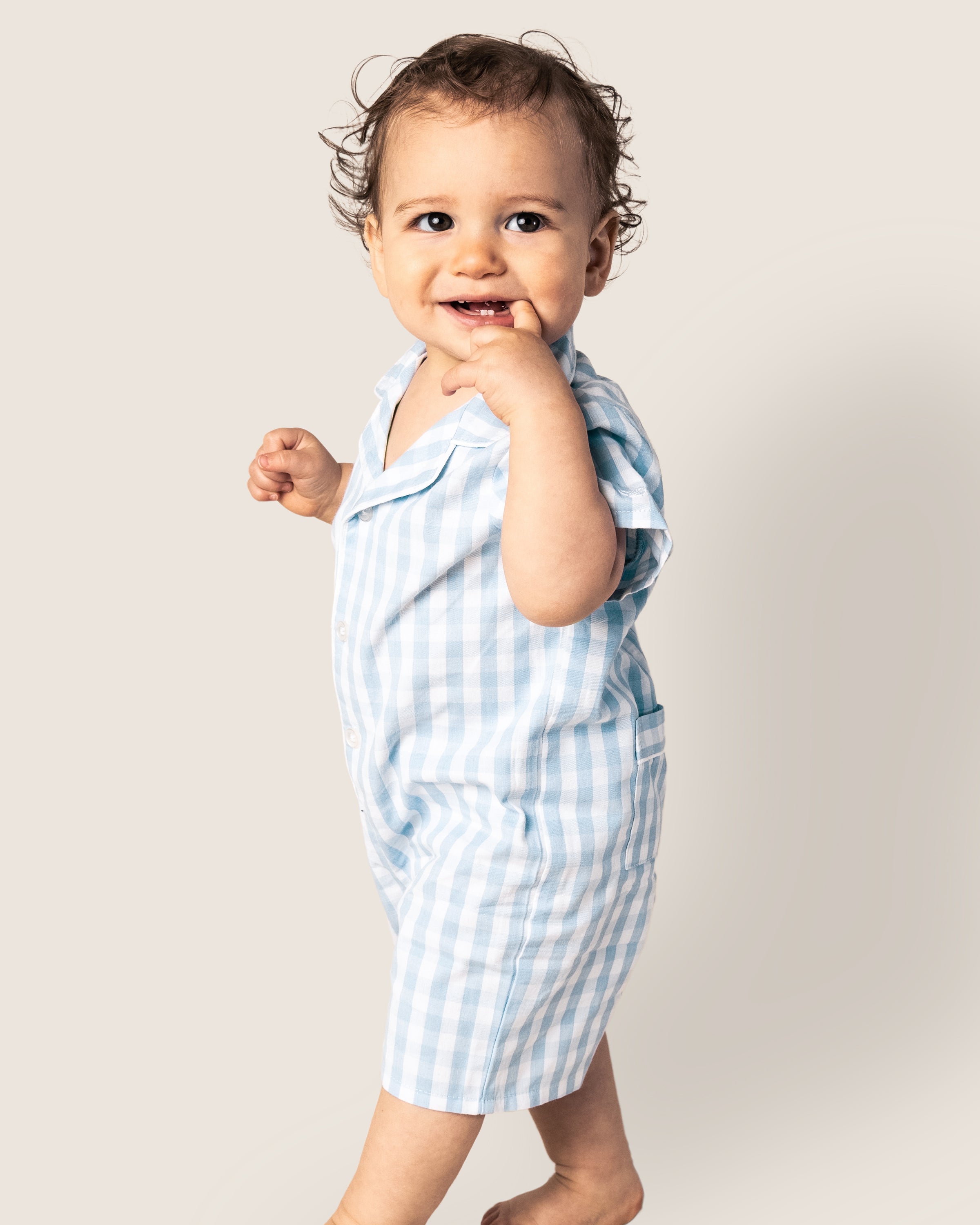 A smiling toddler with curly hair stands barefoot in Petite Plumes Babys Twill Summer Romper, featuring a light blue gingham pattern. One hand is near their mouth as they look at the camera against a plain background.