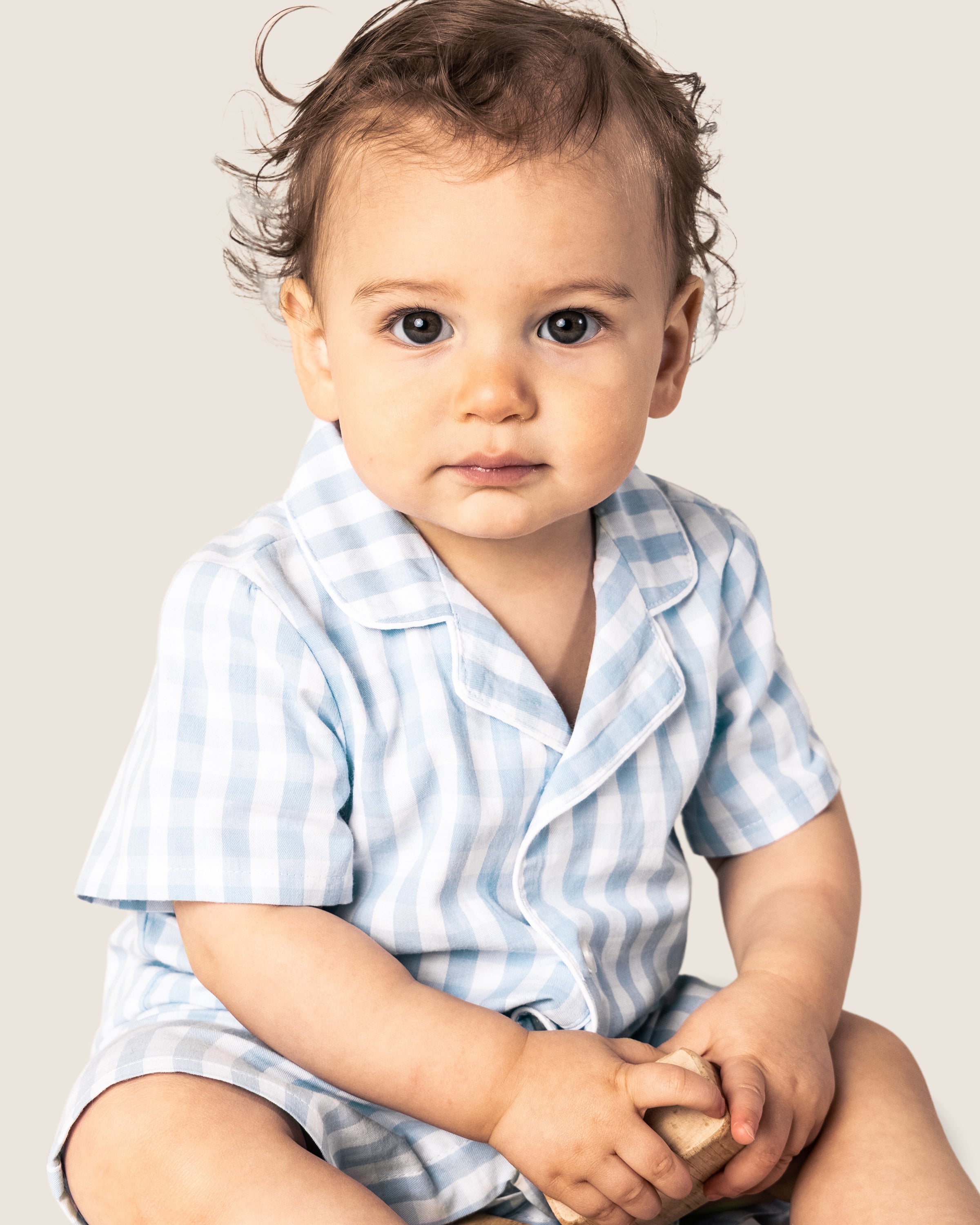 A baby with curly hair wearing Petite Plumes Babys Twill Summer Romper in Light Blue Gingham holds a small object, staring ahead neutrally against a plain, light backdrop.