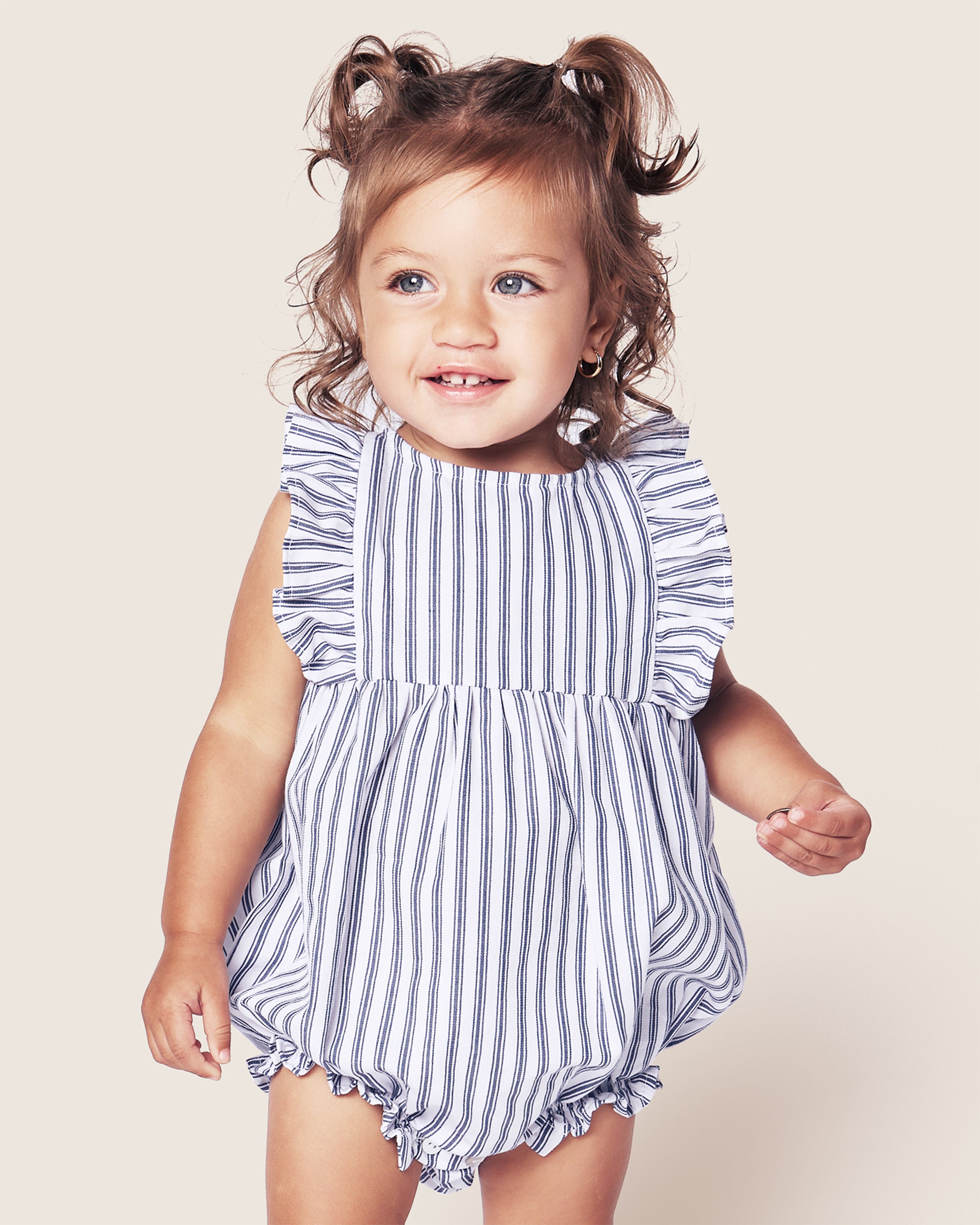 A toddler in Petite Plumes Babys Twill Ruffled Rompers in Navy French Ticking beams against a plain background, her hair styled in adorable pigtails.