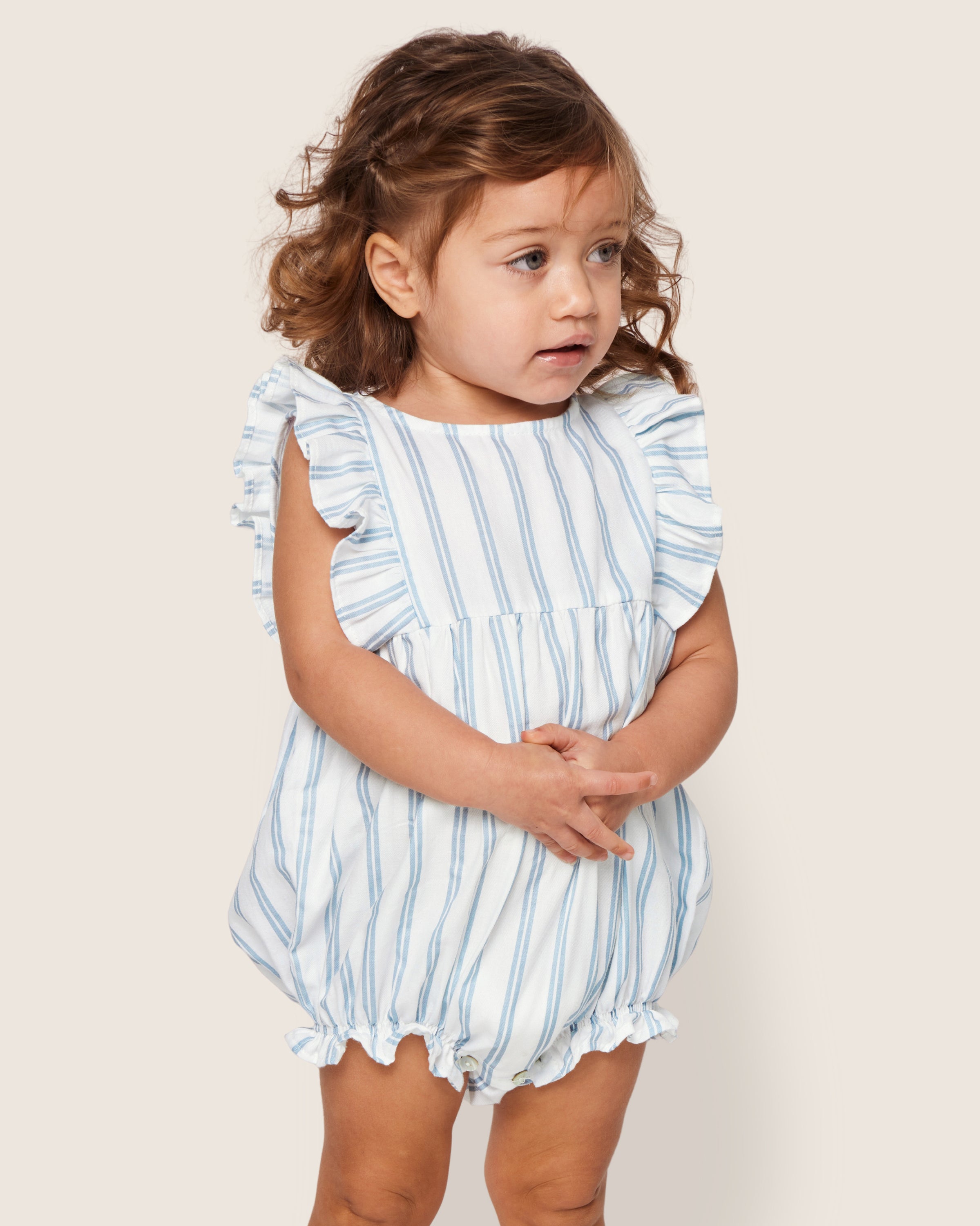 A toddler with curly brown hair stands against a light background, wearing Petite Plumes Babys Twill Ruffled Romper in Periwinkle Stripe. The romper features ruffled sleeves and leg openings, reminiscent of Victorian-era sleepwear. They gaze slightly to the side with hands clasped.