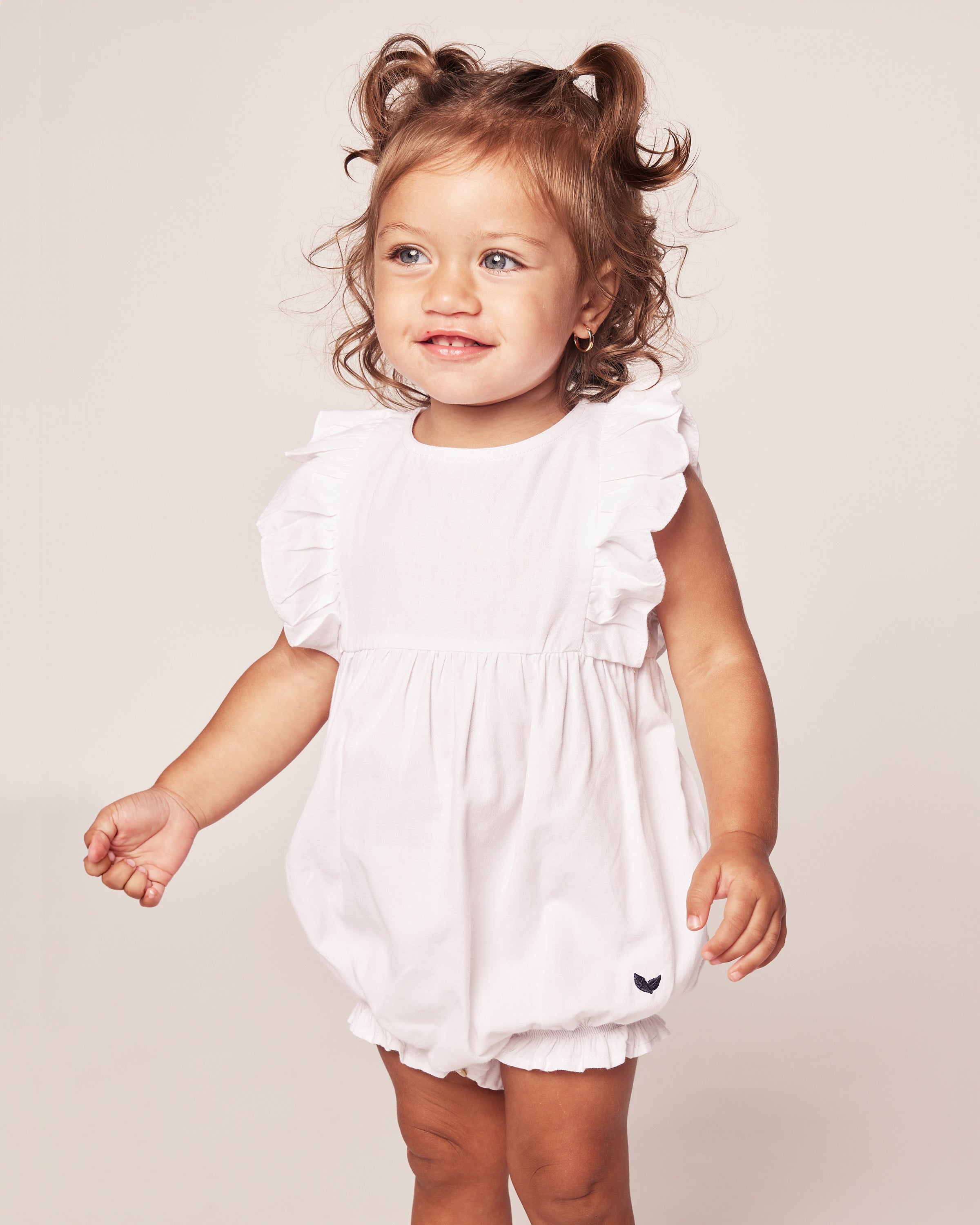 A toddler with curly brown hair tied in pigtails is wearing Petite Plumes Babys Twill Ruffled Romper in white, featuring a small dark heart logo. The child is smiling against a plain background.