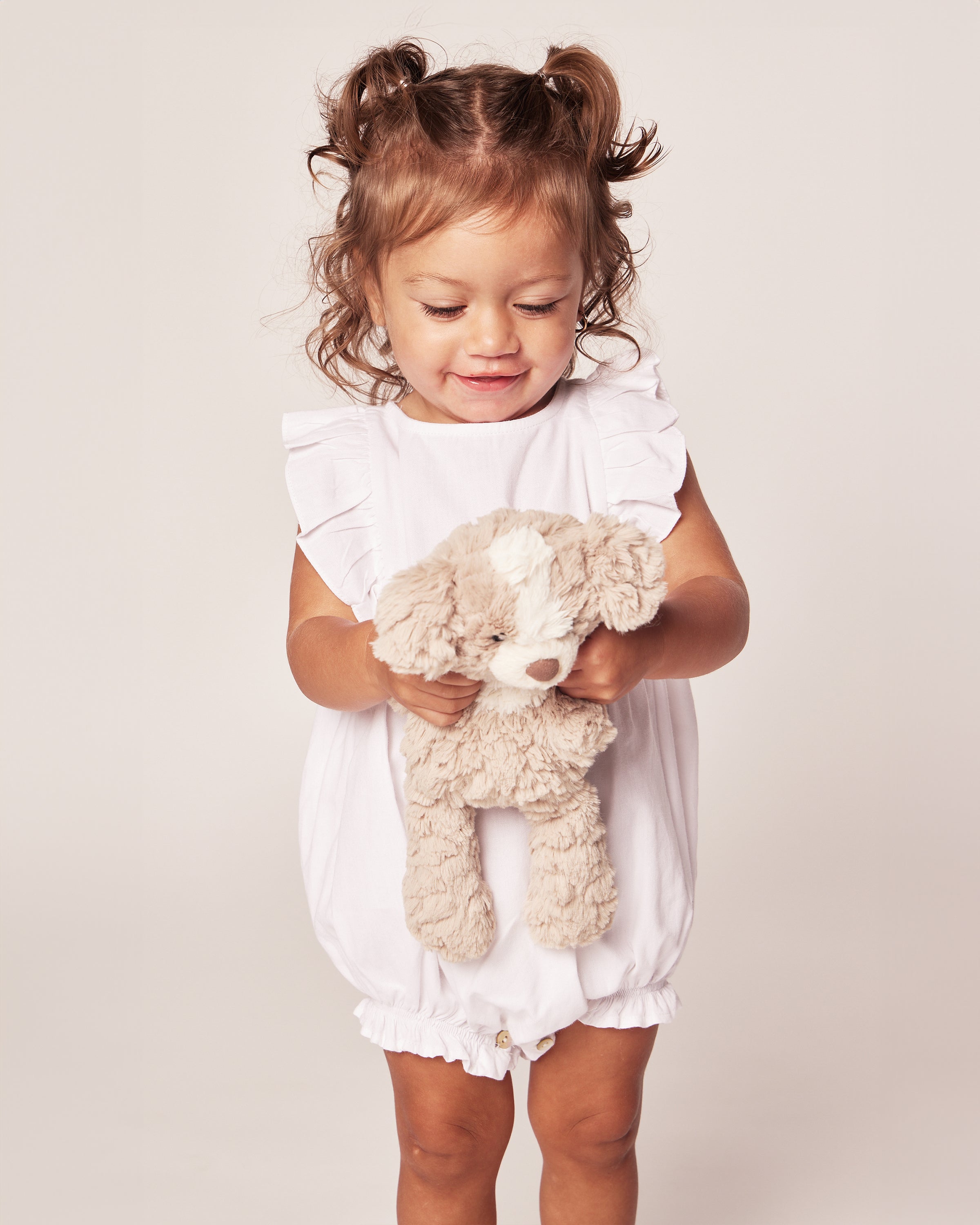 A young child with curly hair smiles while holding a plush dog toy, dressed in Petite Plumes Babys Twill Ruffled Romper in White. The light-colored background enhances the charm of this cozy sleepwear moment.
