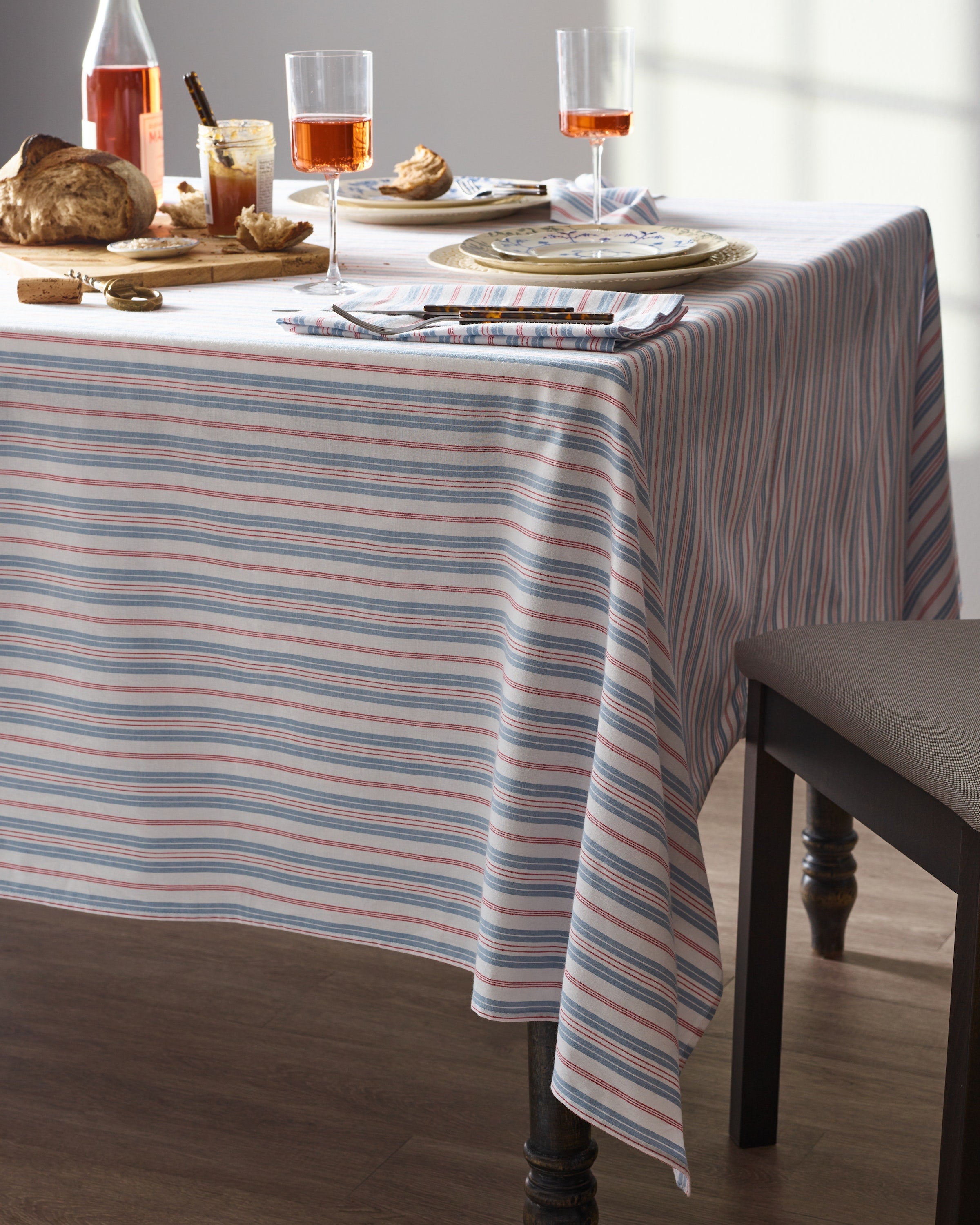 A Signature Twill Tablecloth in Vintage French Stripes by Petite Plume graces the table, accompanied by two glasses of an orange beverage, a bottle, bread, and small dishes. Sunlight filters through the window, casting soft shadows on the wall for a cozy atmosphere.