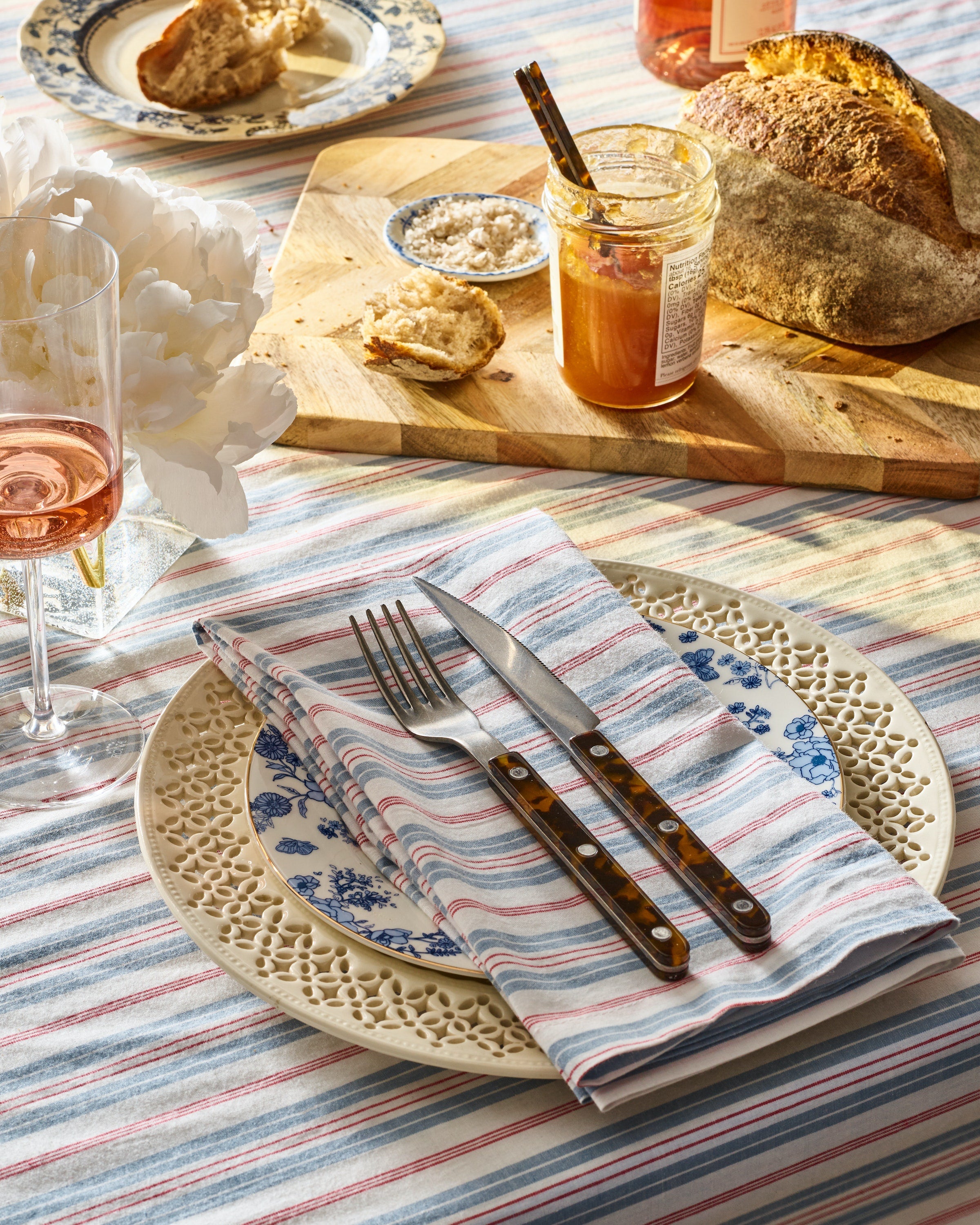 A table features 100% cotton linens with Petite Plume prints, a decorative plate, the Signature Twill Napkins in Vintage French Stripes, fork, and knife. A wooden board holds bread with honey and butter. Completing the look are a glass of pink beverage and a white flower on an elegant striped tablecloth.