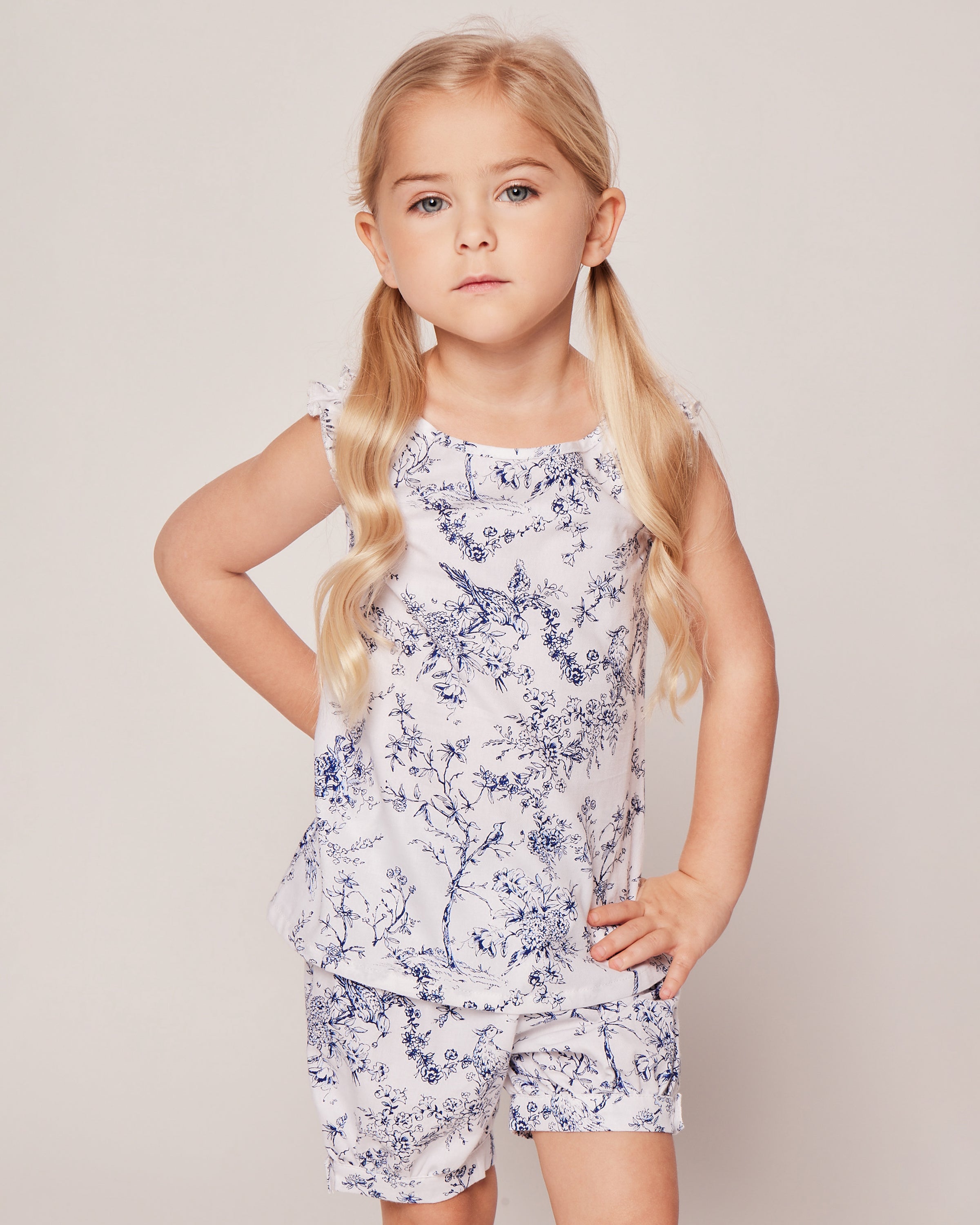A young child with long blonde pigtails stands confidently, wearing the Girls Twill Amelie Short Set in Timeless Toile from Petite Plume. The white sleeveless top and shorts are made of luxury cotton with a blue floral pattern set against a plain background, looking at the camera.