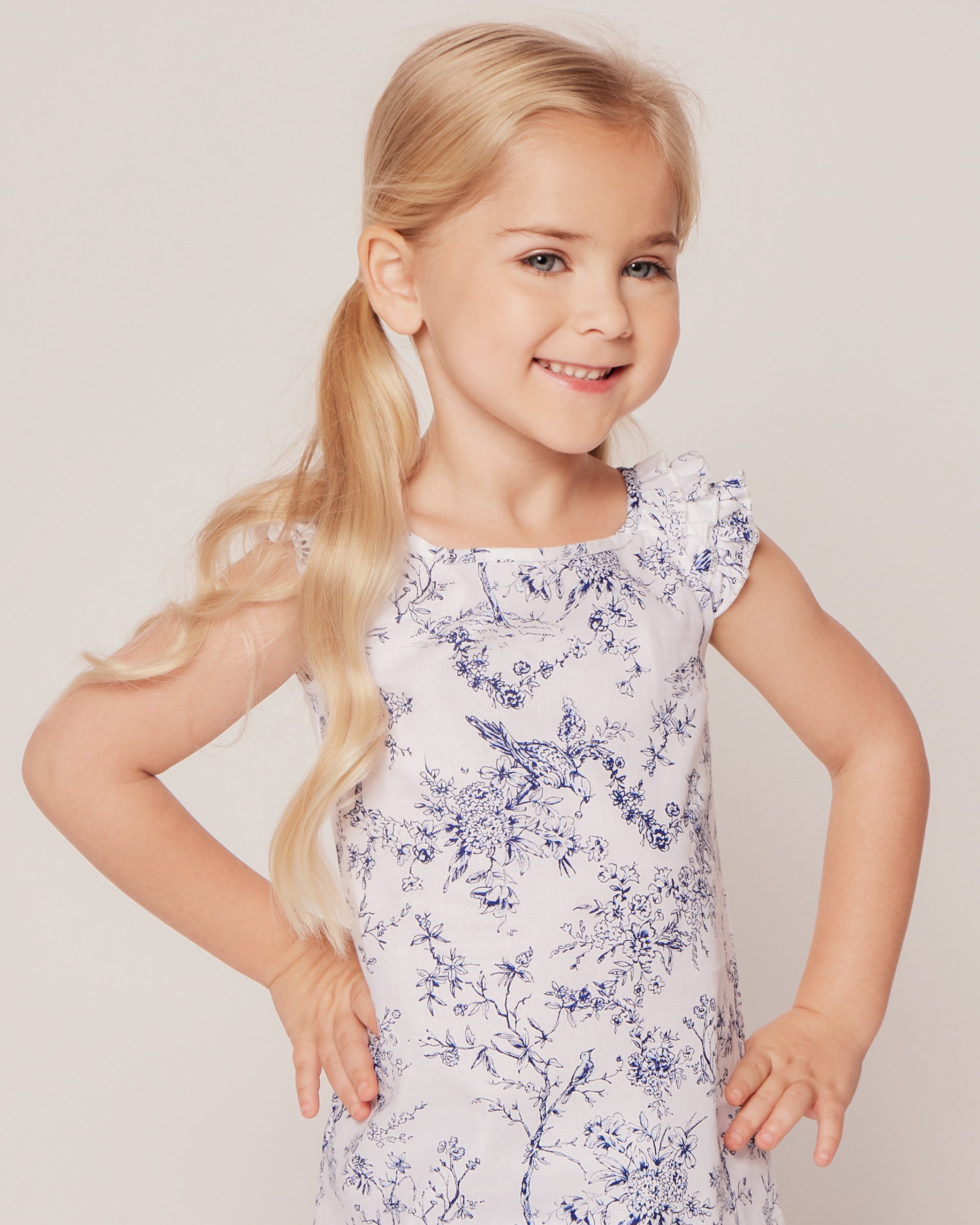 A young girl with long blonde hair smiles, wearing Petite Plumes Girls Twill Amelie Short Set in Timeless Toile. Her hands rest on her hips as she stands against a plain light background, exuding a charming blend of innocence and style.