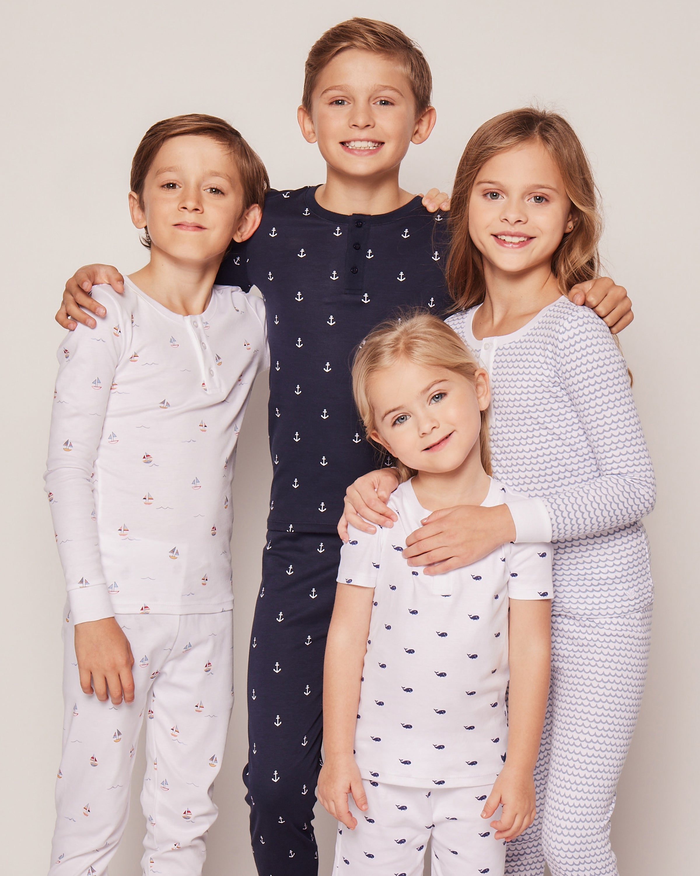 Four children, dressed in matching Petite Plume Kids Pima Snug Fit Pajama Sets in Portsmouth Anchors, smile together. The outfits feature anchor and wave patterns in white and navy blue on a light background, crafted from soft Pima Cotton with long sleeve tops and pants.