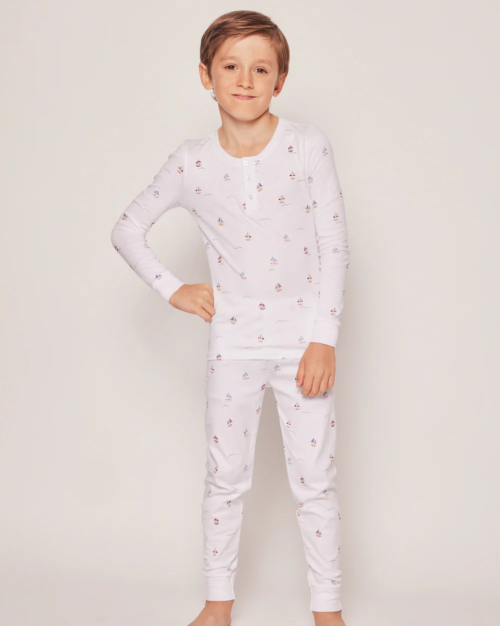 A young boy, barefoot and adorned in Petite Plumes Boys Pima Snug Fit Pajama Trio with small sailboat prints, stands against a plain backdrop. Crafted from soft Pima cotton, his pajamas promise comfort and sweetness as he smiles gently with hands at his sides.