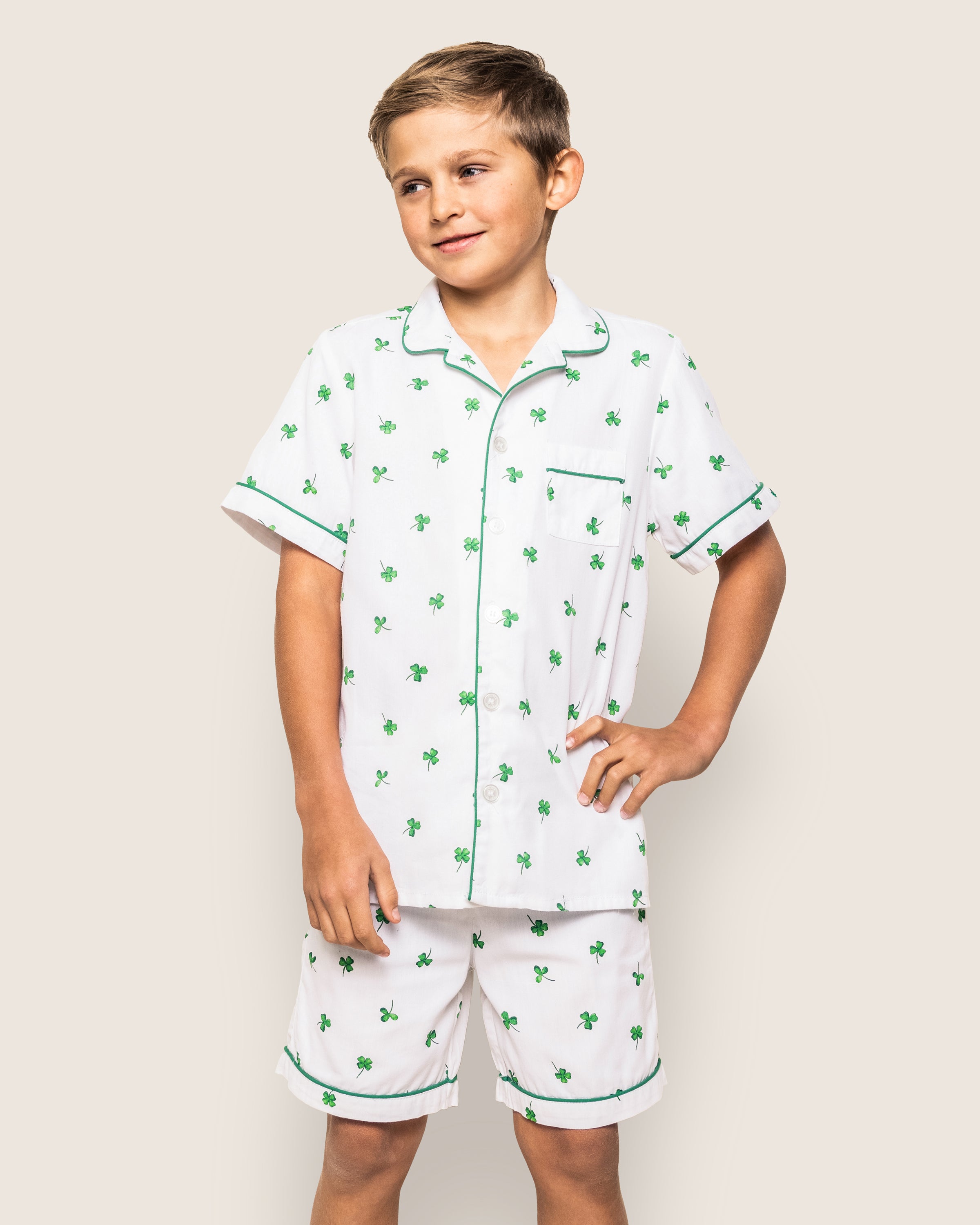 A child models the Kids Twill Pajama Short Set in Shamrocks from Petite Plume, standing against an off-white background. The white short-sleeve pajamas have green clovers and dark piping, with a hand in their pocket and a smile completing the Classic Short Set look.