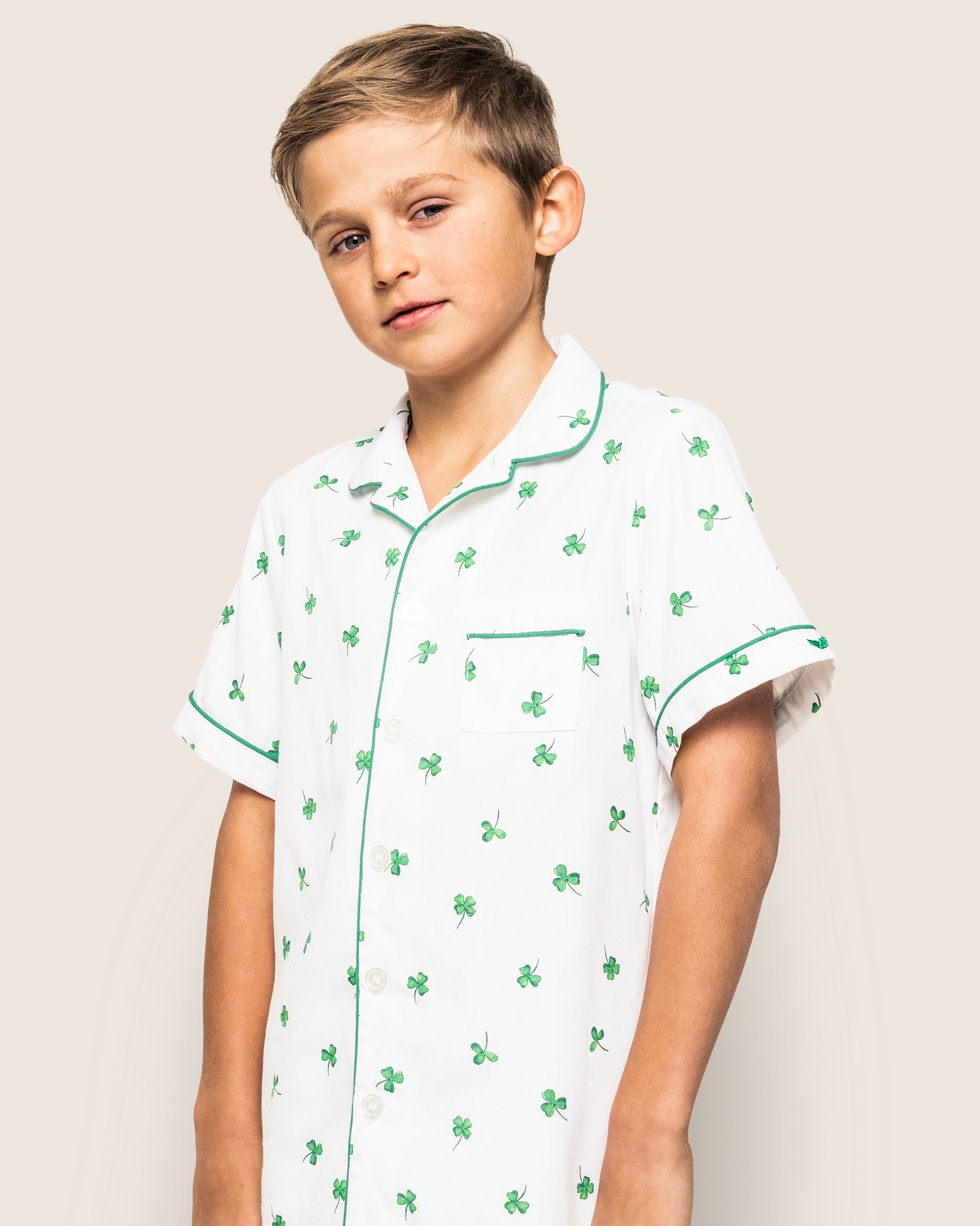 The young boy wears a Petite Plume Kids Twill Pajama Short Set in Shamrocks, featuring green clover patterns, a chest pocket, and green trim. He poses against a light background, looking slightly to the side.