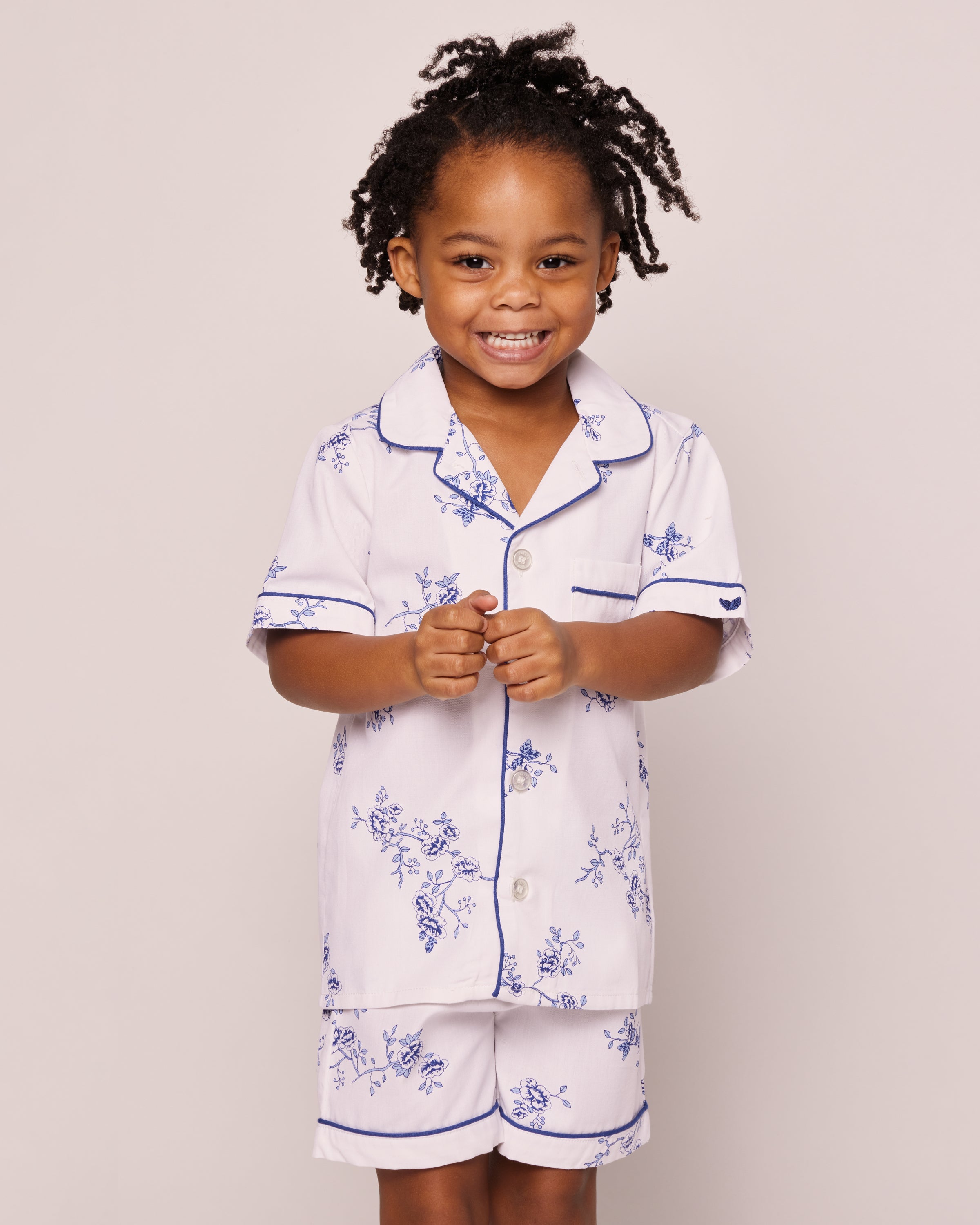 A child smiles in Petite Plumes Kids Twill Pajama Short Set in Indigo Floral, featuring a luxurious short-sleeved, collared top with buttons and matching shorts made of premium cotton. Their hair is styled in twists, adding to the cheerful vibe against the plain backdrop.