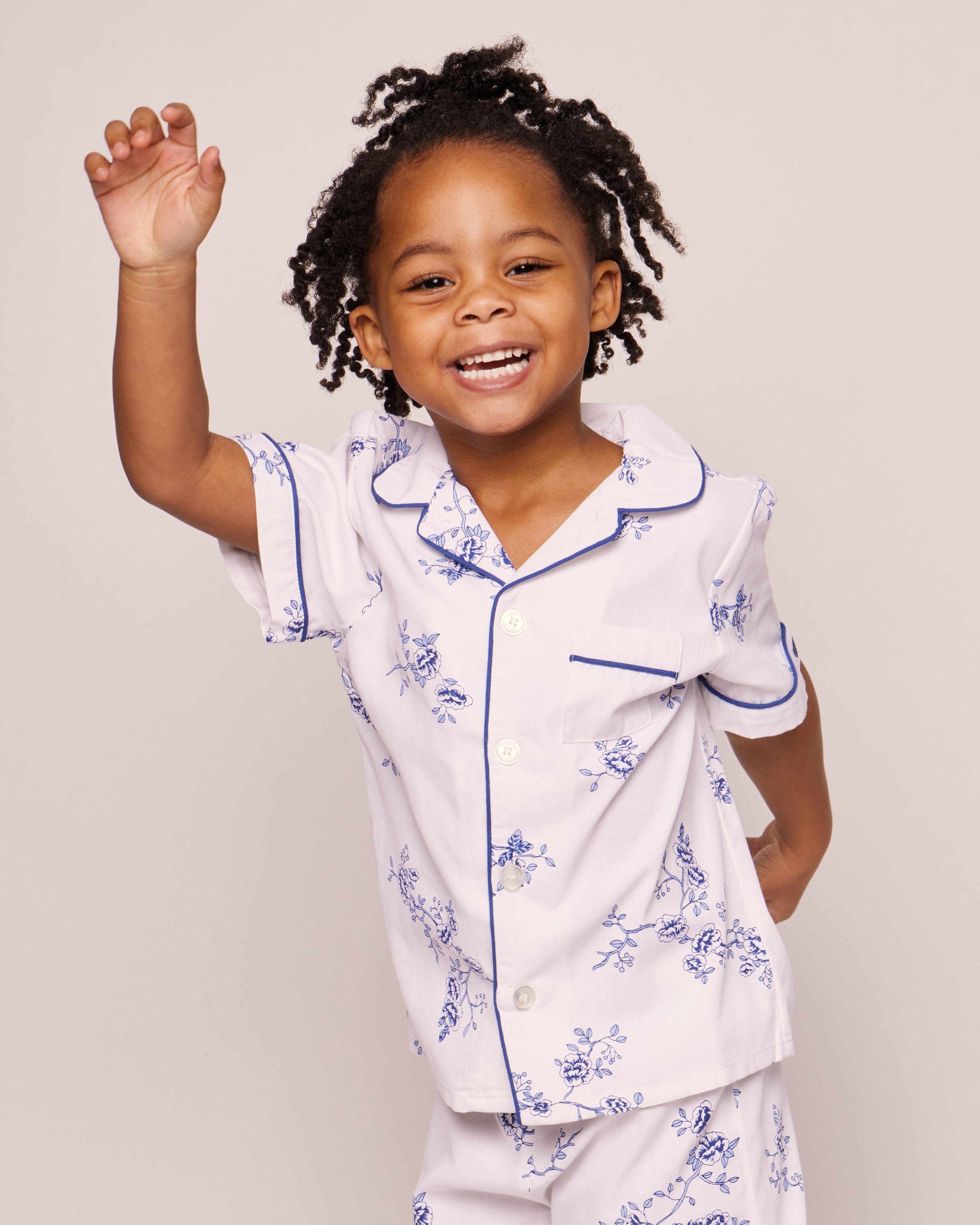 A smiling young child with curly hair raises one hand while wearing Petite Plumes Kids Twill Pajama Short Set in Indigo Floral, featuring the finest quality white cotton with blue floral patterns and blue-trimmed collar and pocket, set against a plain neutral background.