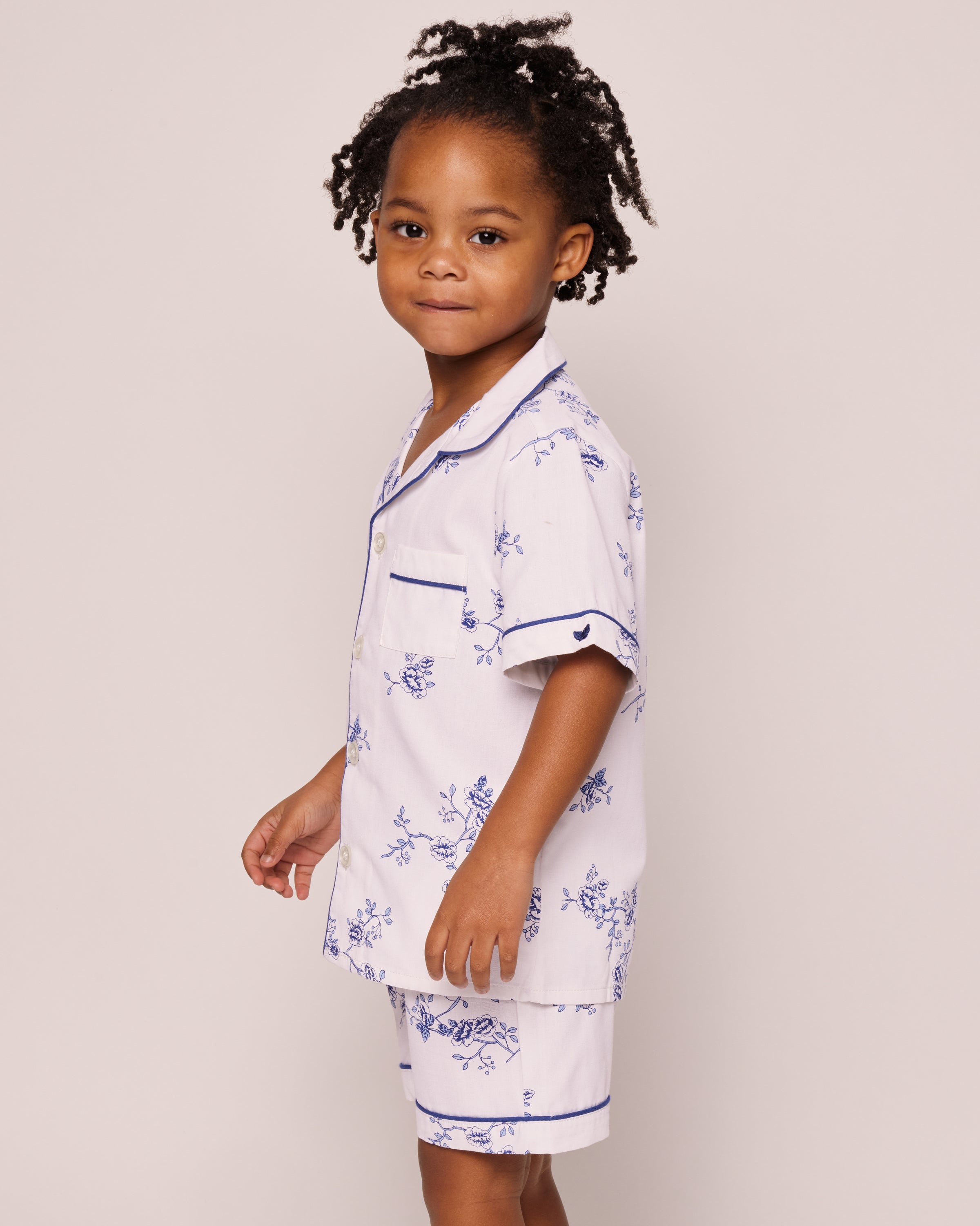 A young child with curly hair wears Petite Plumes Kids Twill Pajama Short Set in Indigo Floral, featuring white sleepwear with a blue floral design, a pocketed top, and navy piping. The child stands against a plain light background, gazing slightly to the side.