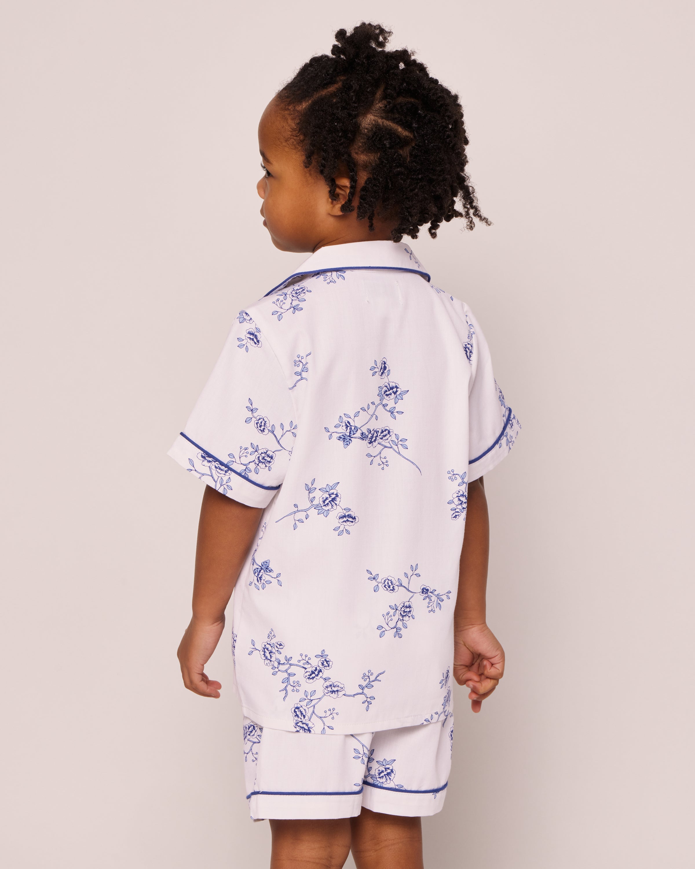 A child stands facing away, curly hair visible, wearing Petite Plumes Kids Twill Pajama Short Set in Indigo Floral. The white, short-sleeved cotton set with a blue floral vintage print is set against a plain, light background.