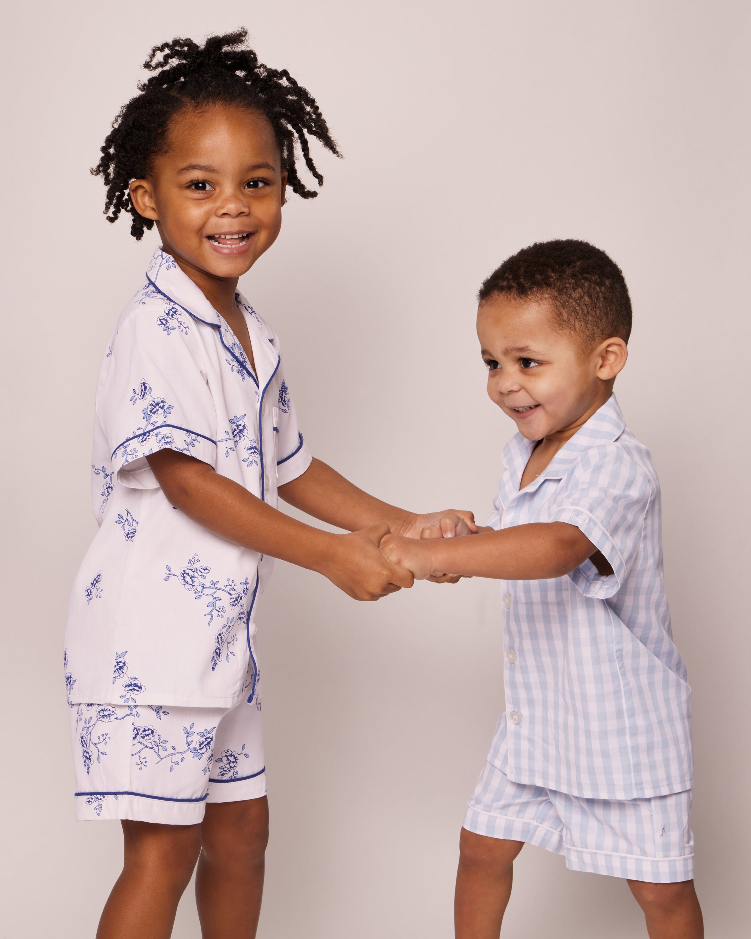 Two young children smile and hold hands in luxurious Petite Plume sleepwear. The child on the left wears the Kids Twill Pajama Short Set in Indigo Floral made of high-quality cotton, while the child on the right sports a light blue and white checkered set. Neutral background.