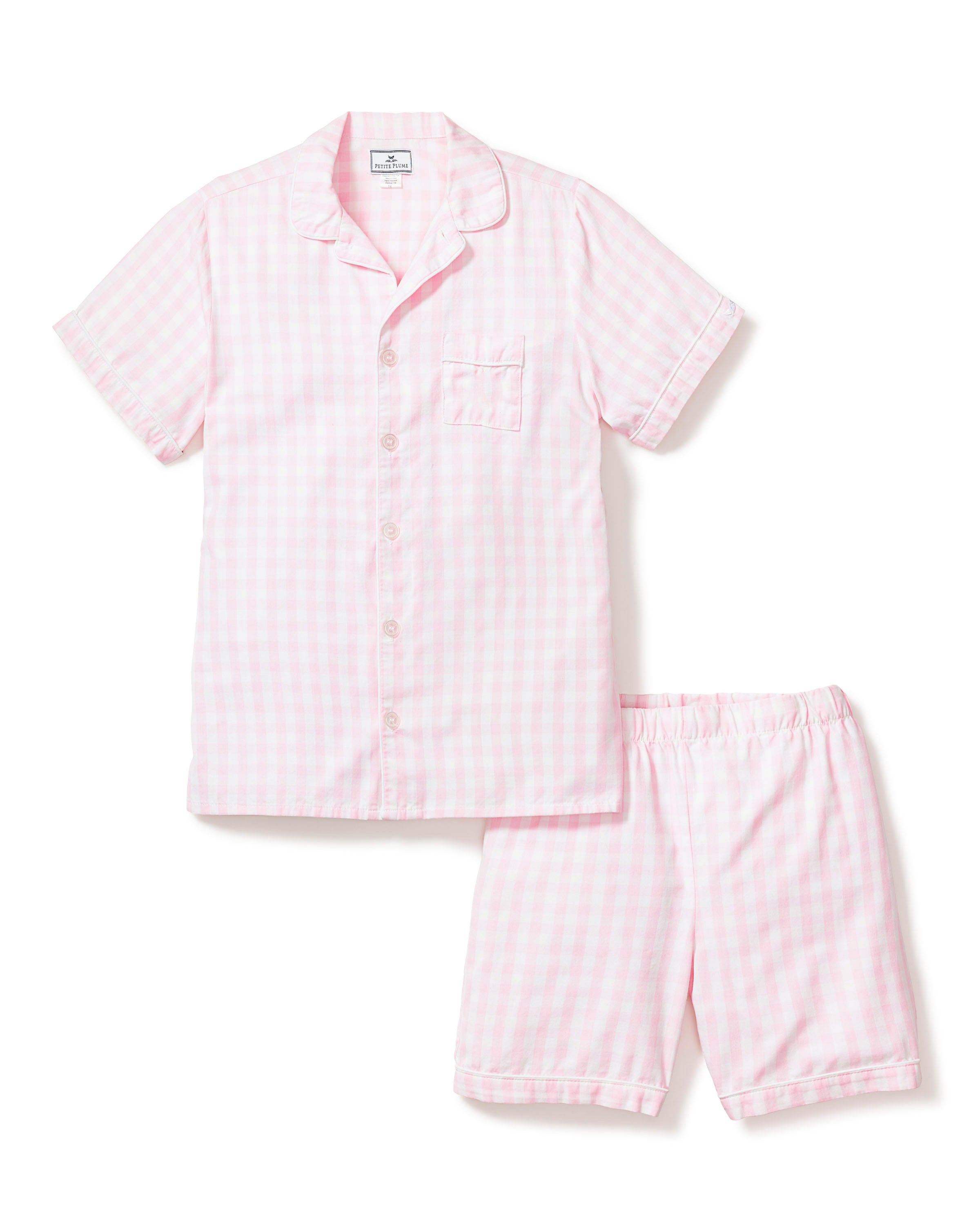 Three folded Petite Plume Kids Twill Pajama Short Sets in pink gingham rest on a textured surface, featuring charming designs of pink hearts, floral toile patterns, and pink stripes. Crafted from yarn-dyed cotton, they exude quality. Elegantly surrounded by white flowers for enhanced presentation.