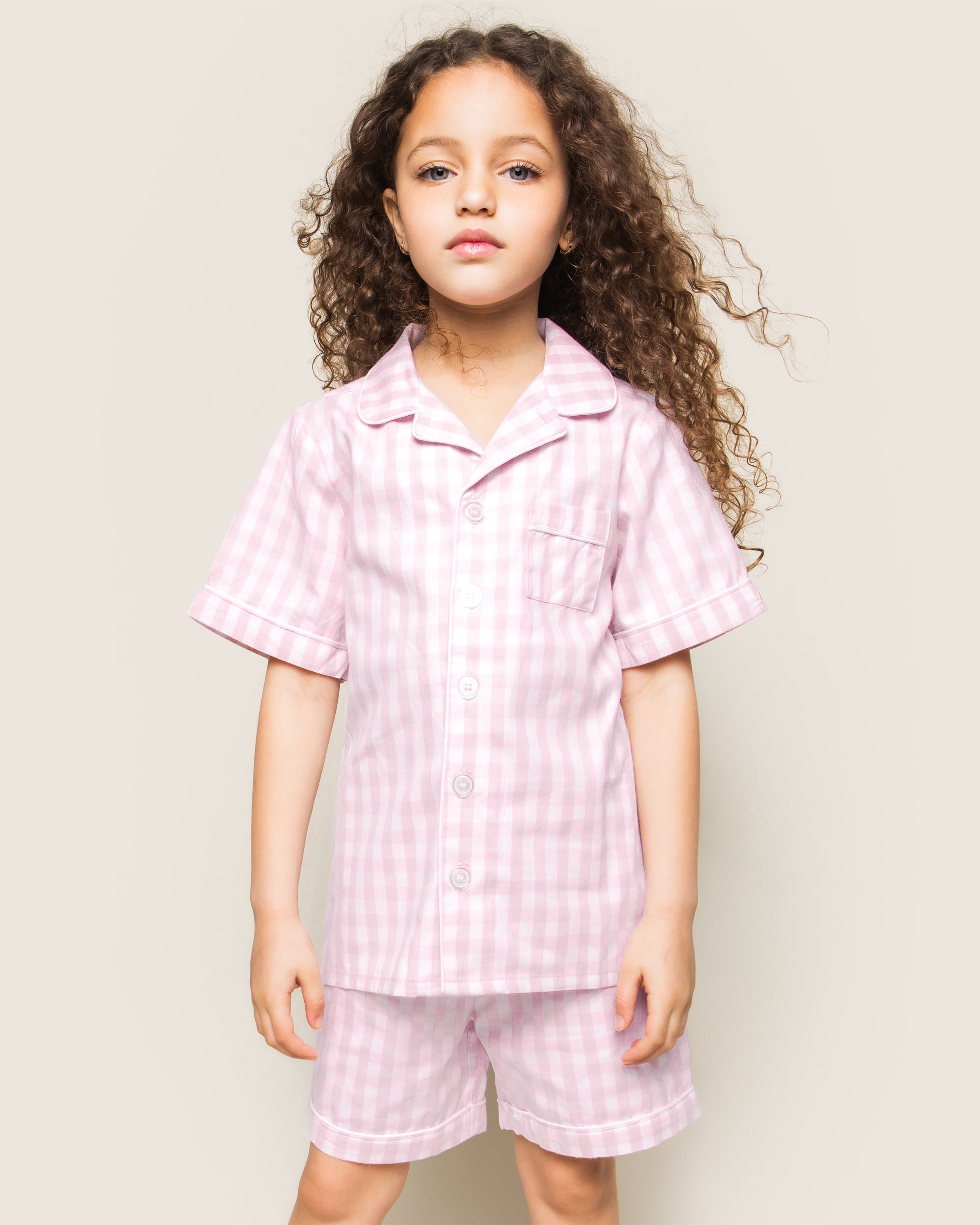 A curly-haired child is wearing the Petite Plume Twill Pajama Short Set in pink gingham, standing against a plain background.