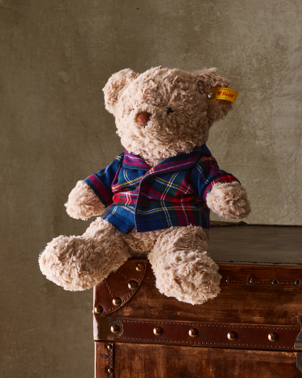 A Petite Plume x Steiff Teddy Bear in Windsor Tartan, wearing a blue, red, and green plaid collared pajama top, sits on a vintage-style trunk.