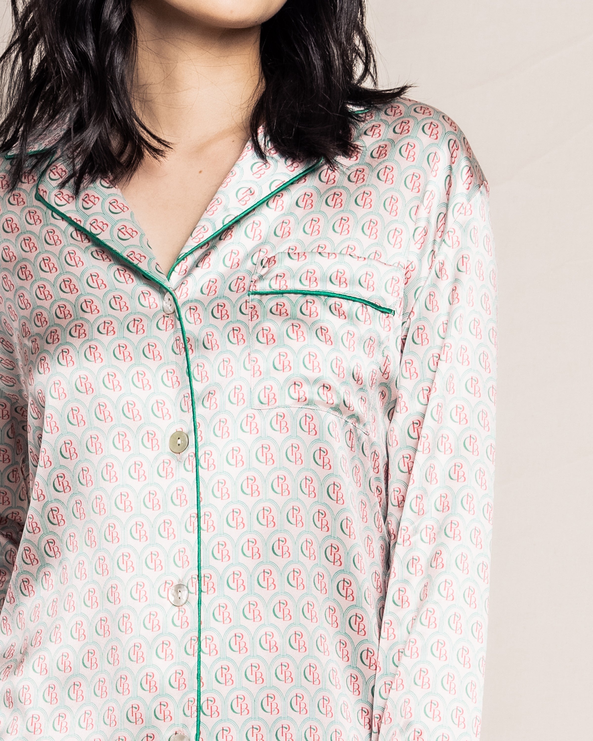 The woman is wearing the cream Petite Plume Womens Silk Pajama Top from the exclusive Colony Hotel collection, showcasing a charming pink and red pattern with green piping. The image shows her from the neck down, with hair softly falling on her shoulders.