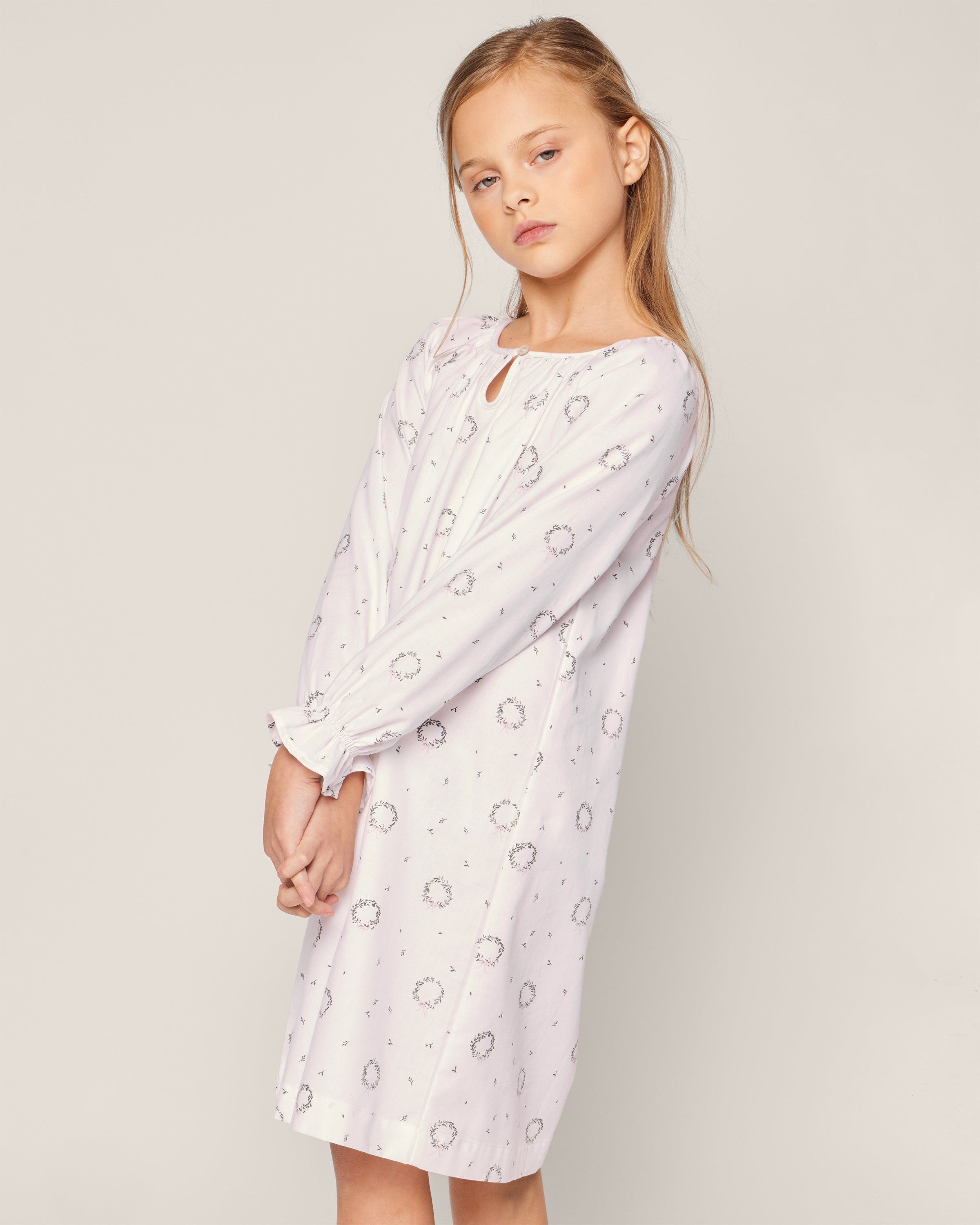 A young girl with long hair wears the Petite Plume Girls Twill Delphine Nightgown in Somerset Wreath—a light-colored, long-sleeved gown with subtle patterns made from chemical-free fabric. She stands against a plain background, looking slightly to the side.