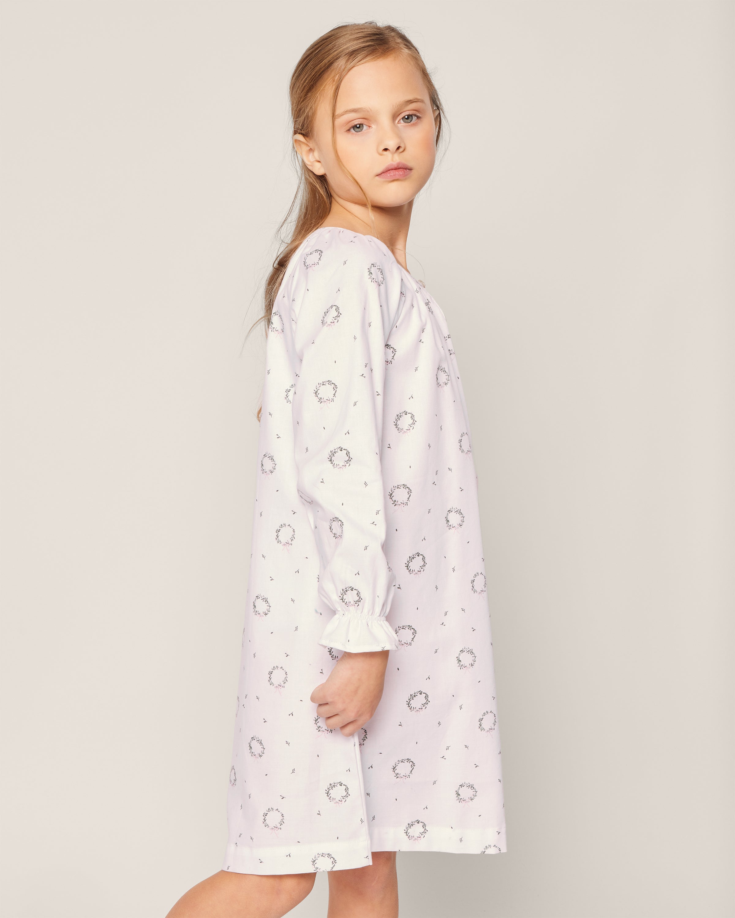 A young girl stands sideways wearing Petite Plumes Girls Twill Delphine Nightgown in Somerset Wreath, light purple with a subtle floral pattern. Her long, light brown hair and calm expression are set against a plain, light background.