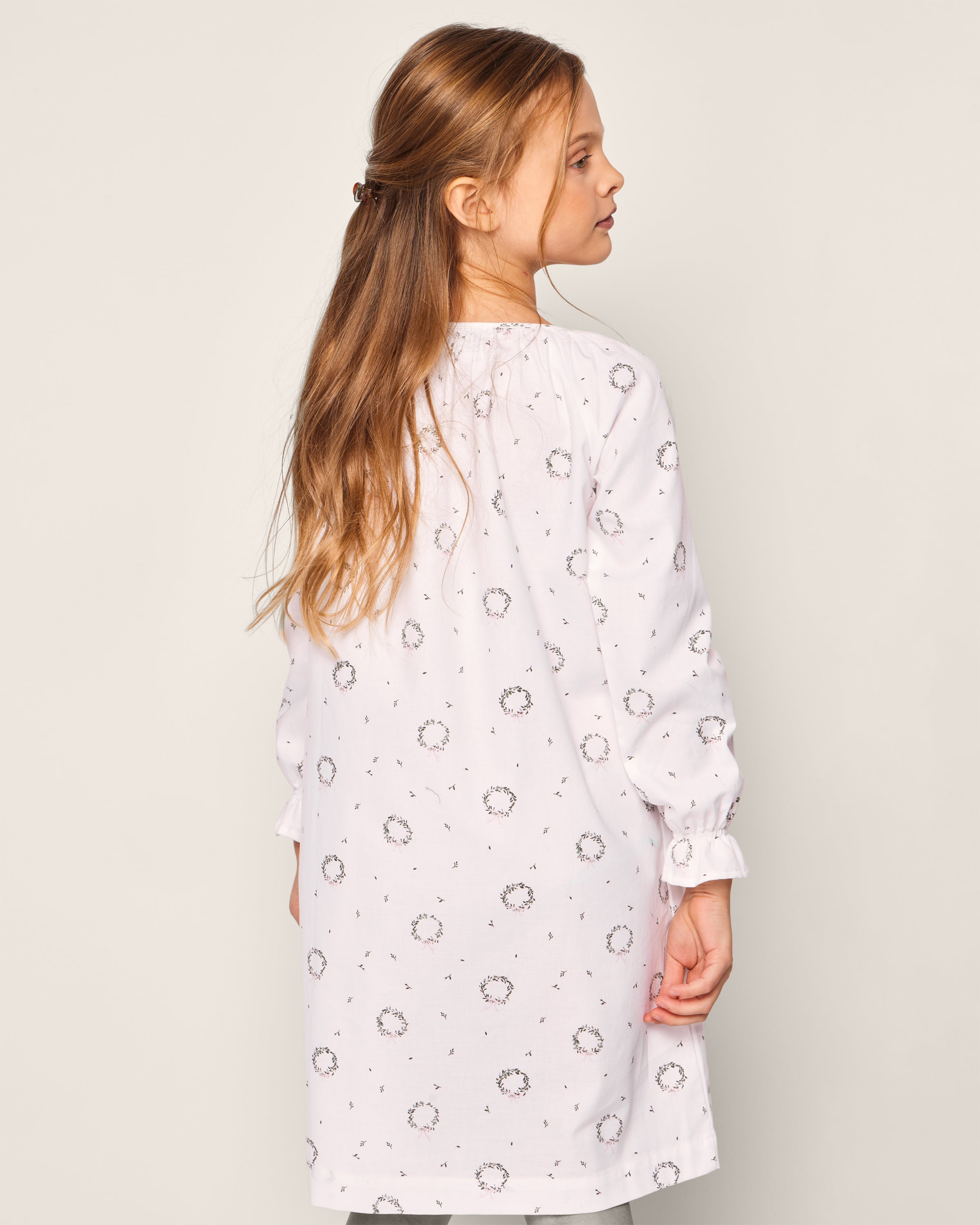 A young girl with long brown hair, partially pulled back, stands slightly turned away in Petite Plumes Girls Twill Delphine Nightgown in Somerset Wreath. The plain background highlights her charming childrens sleepwear.